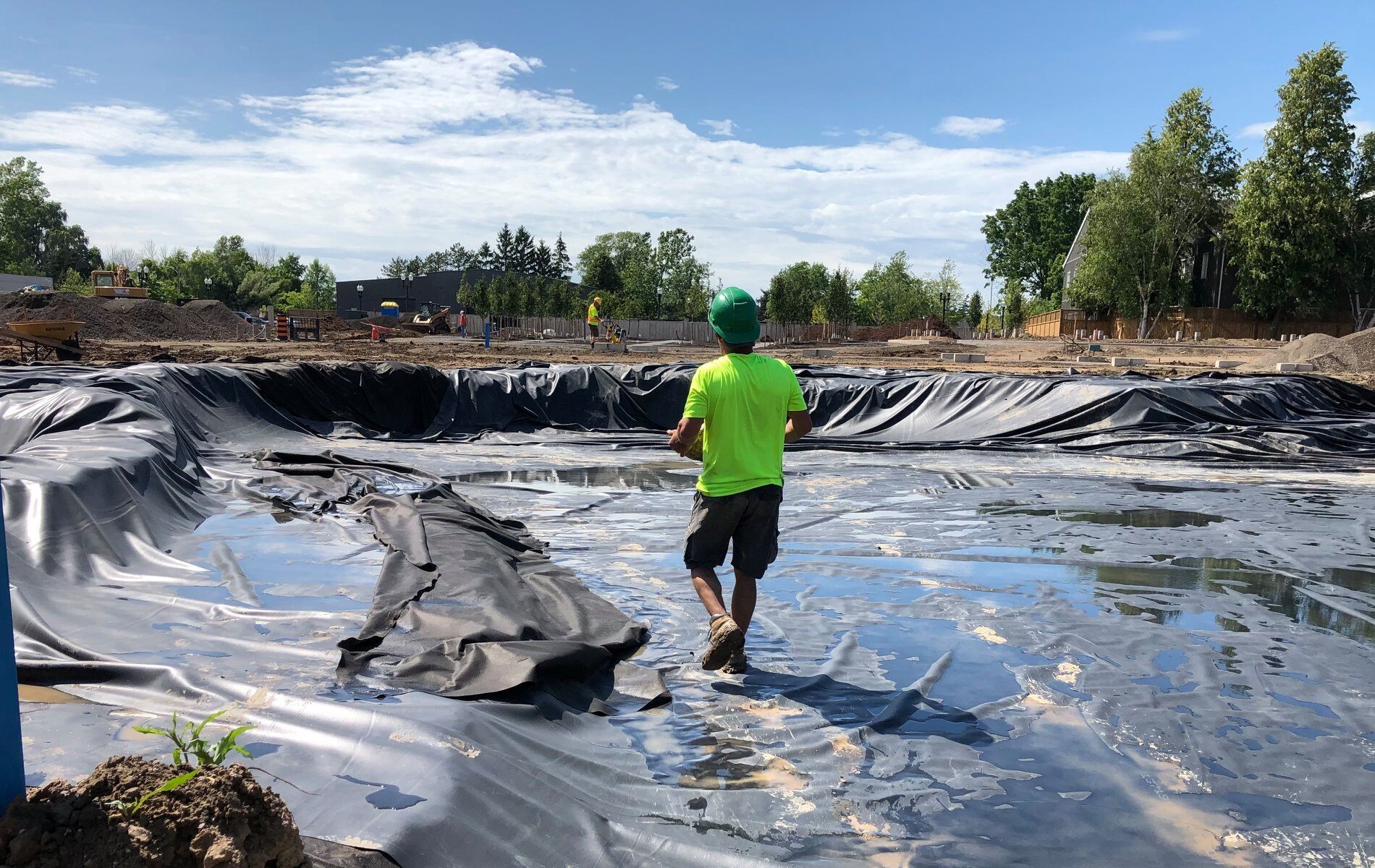 A person in a green shirt and helmet walks across a construction site covered in black plastic, surrounded by trees and machinery.