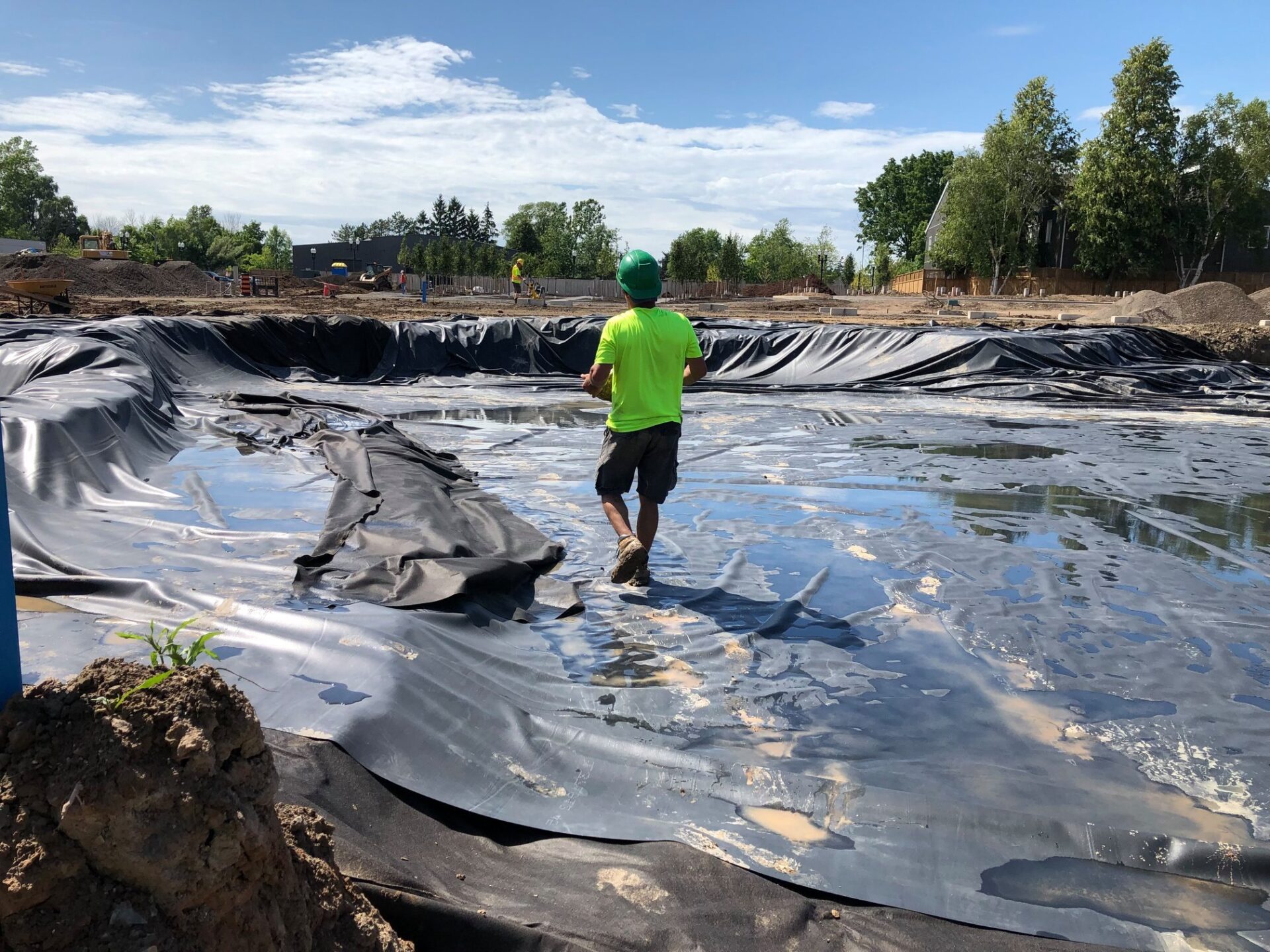 A person in a green shirt and helmet is walking on a large, lined pit at a construction site surrounded by trees.