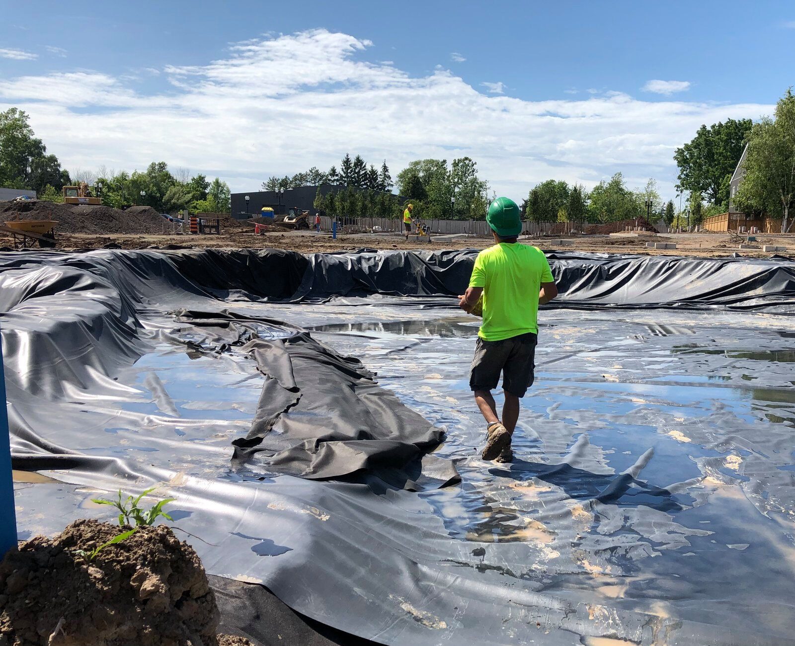 A person in a green shirt and helmet is walking on a large, lined pit at a construction site surrounded by trees.