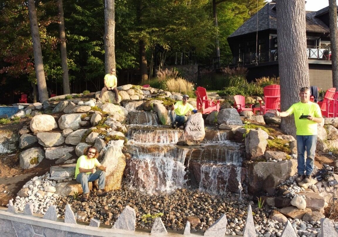 Four persons in bright shirts sit by a rock waterfall. Red chairs and a wooden building are visible in the wooded background.