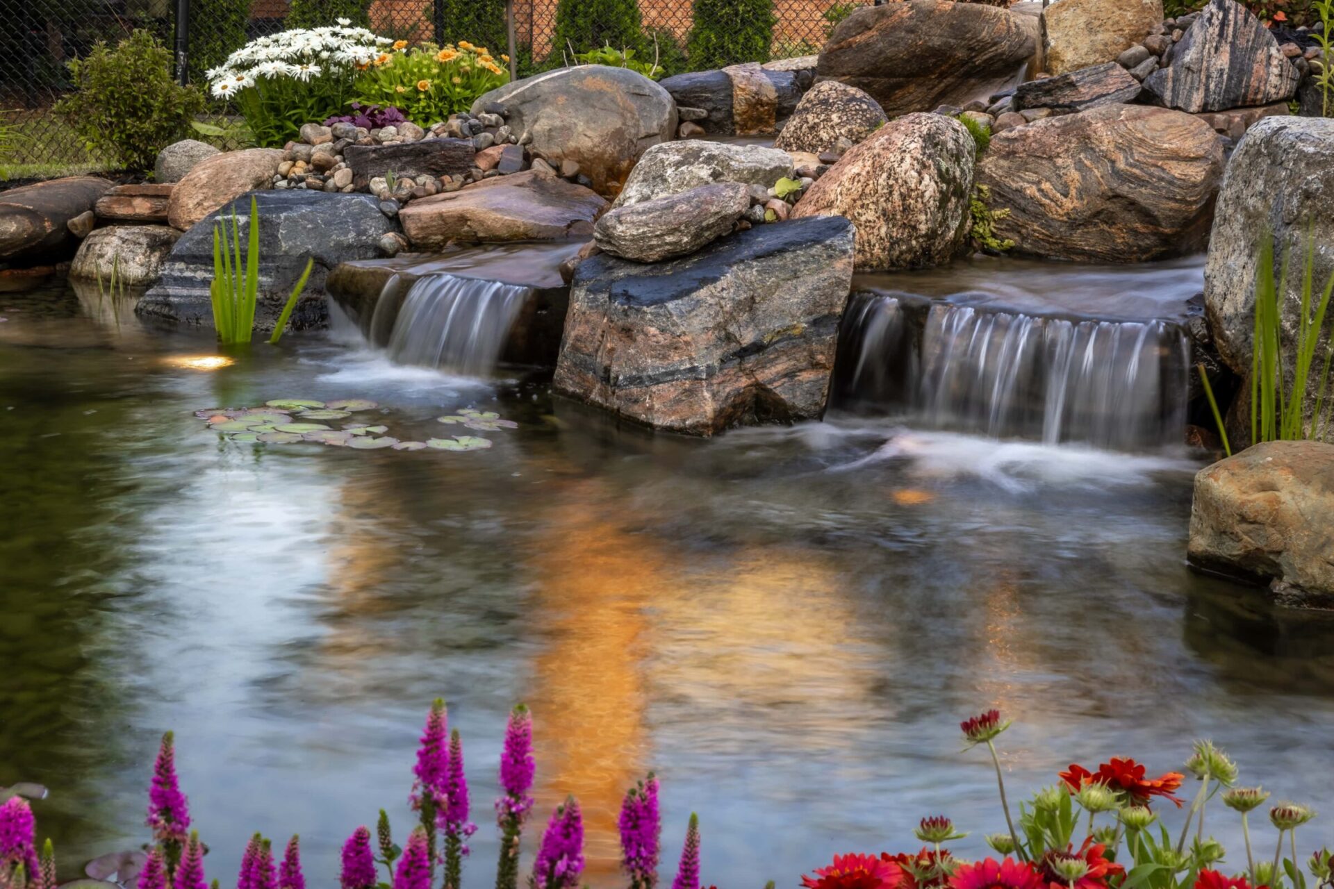 A tranquil garden scene with a small waterfall, surrounded by stones, colorful flowers, and water plants reflecting sunlight in the pond.