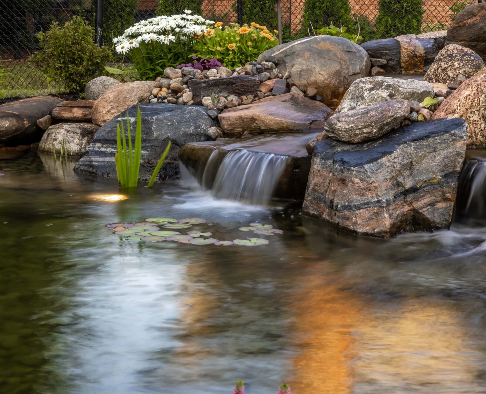 A tranquil garden scene with a small waterfall, surrounded by stones, colorful flowers, and water plants reflecting sunlight in the pond.