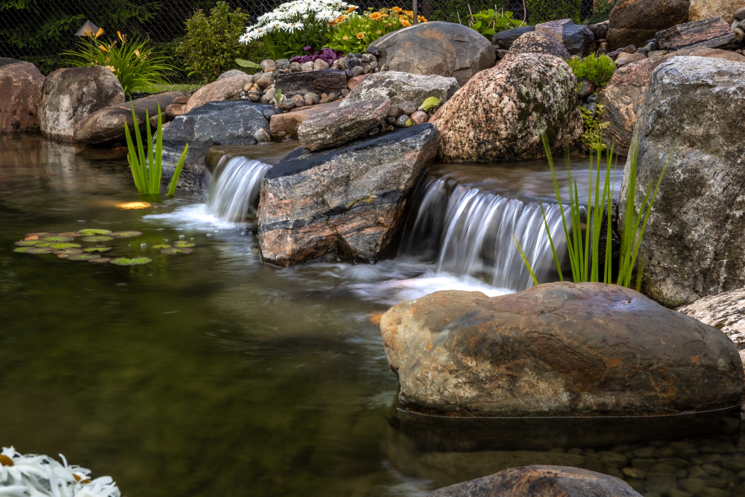 A tranquil garden pond with a small waterfall, surrounded by rocks and plants. Peaceful water flow and green foliage accentuate the scene.