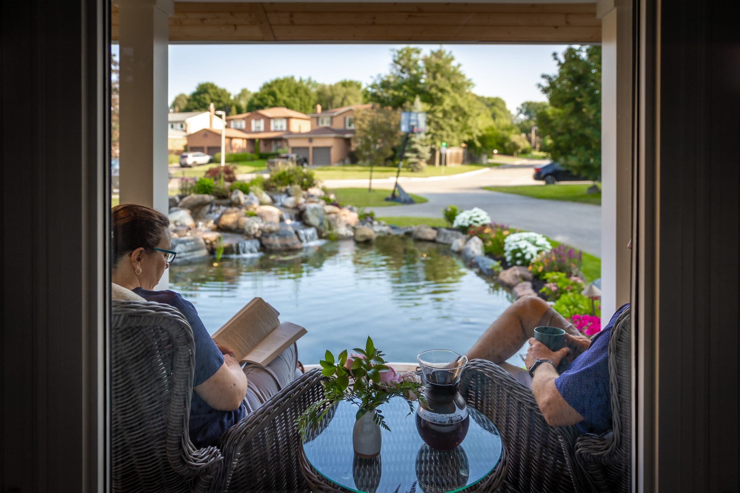 Two people relax by a backyard pond, sitting on wicker chairs, one reading, surrounded by lush greenery and suburban homes in the background.