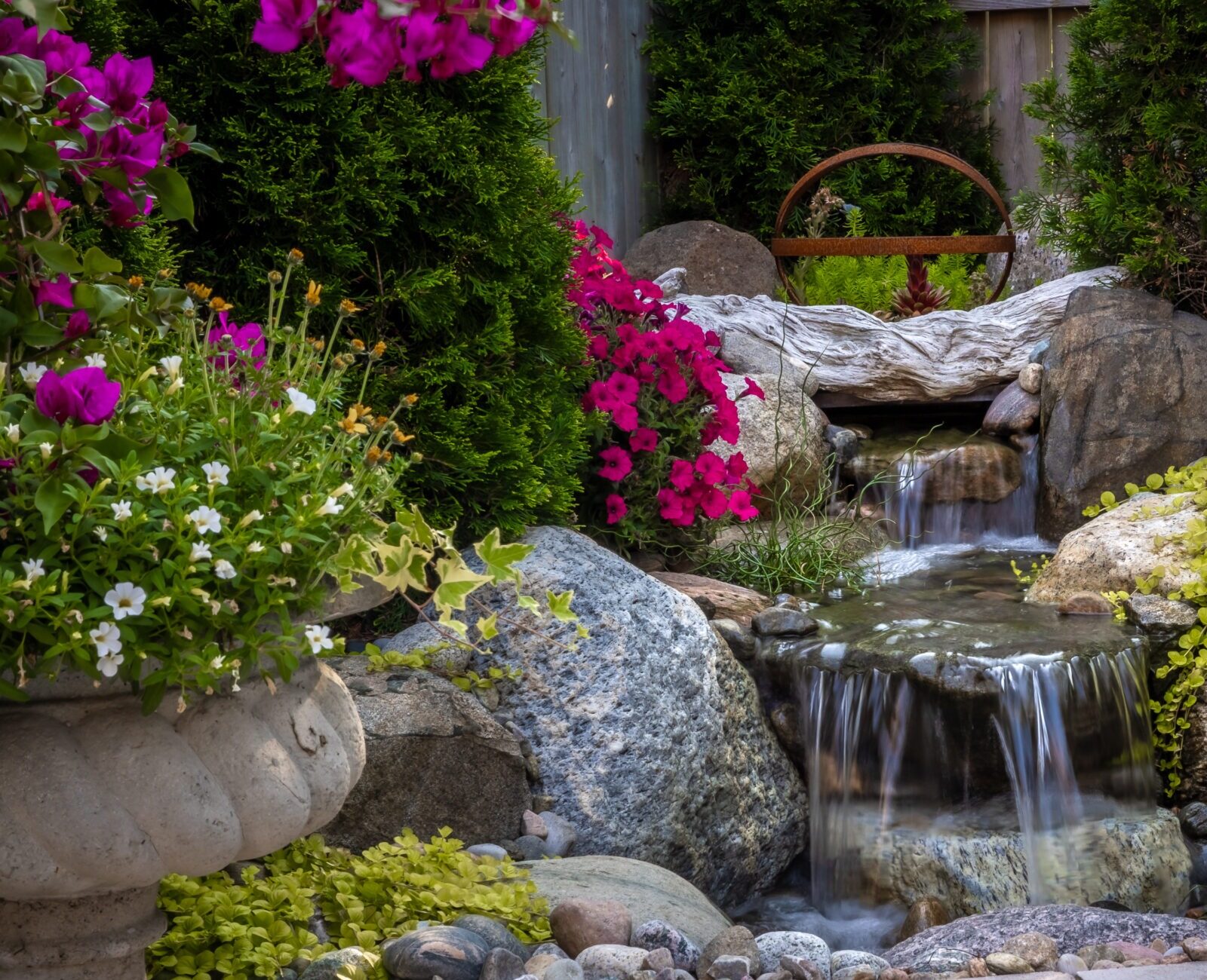 A small garden waterfall cascades over rocks, surrounded by vibrant pink and white flowers, lush greenery, and a decorative stone urn.