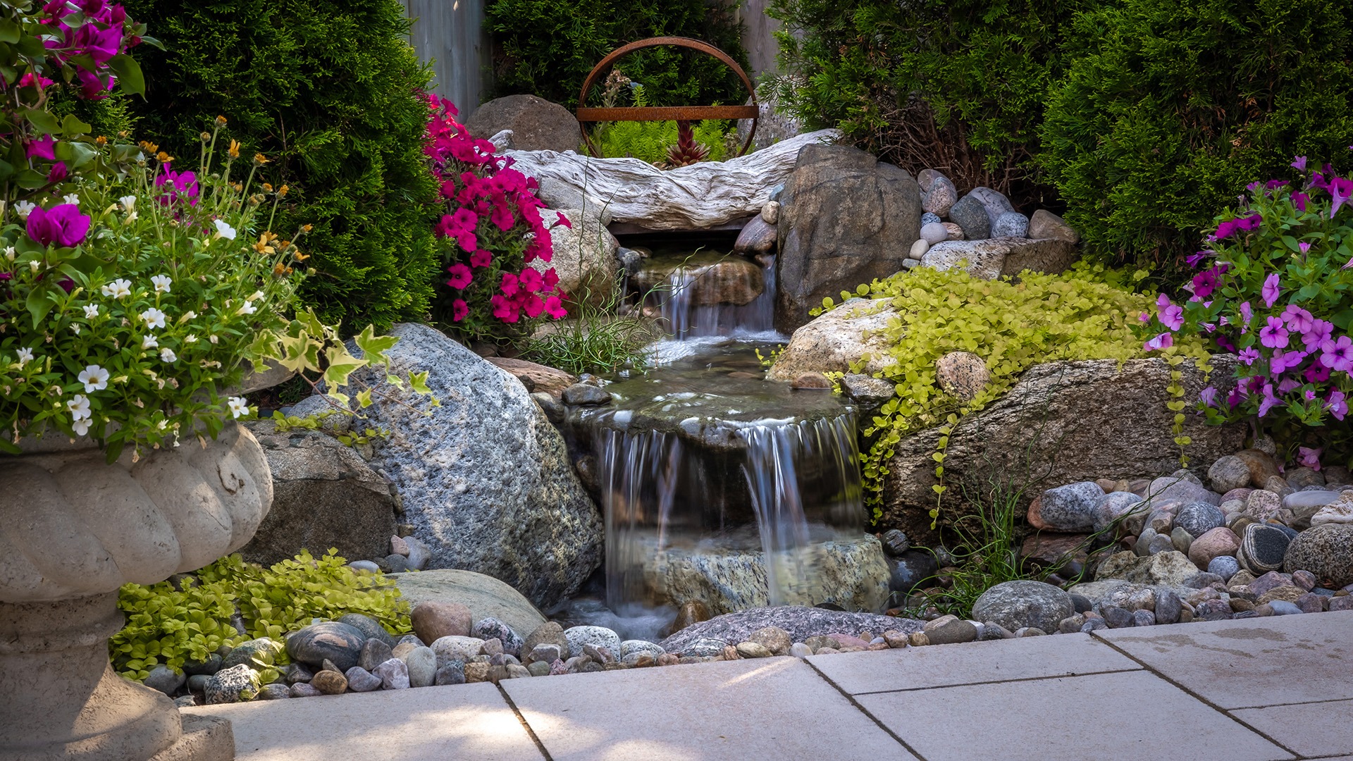 A small garden waterfall flows over rocks, surrounded by vibrant flowers and greenery, creating a serene, natural atmosphere. No people or landmarks.