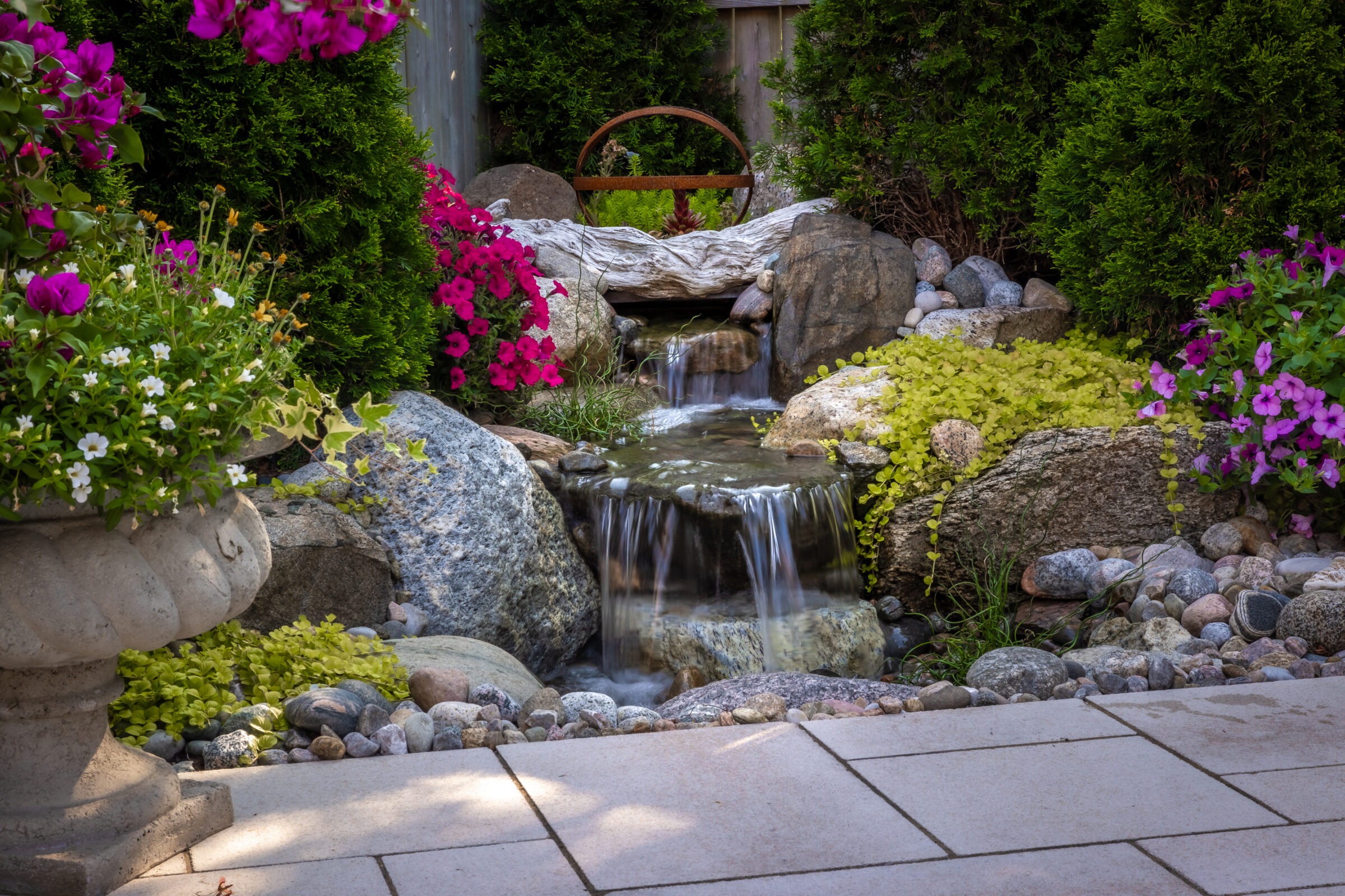 A serene garden scene with a small waterfall, surrounded by rocks, lush greenery, and vibrant purple flowers, creating a tranquil atmosphere.