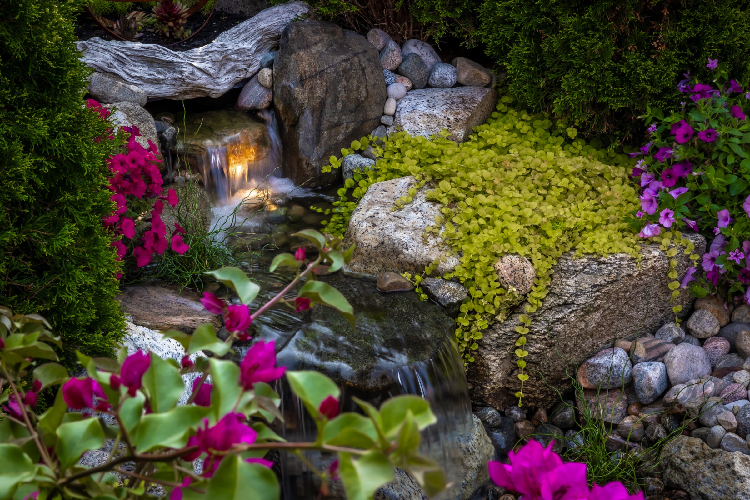 A small, serene garden waterfall surrounded by vibrant flowers, lush greenery, and smooth stones, creating a tranquil natural setting.