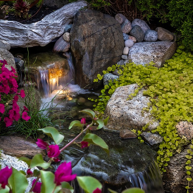 A serene garden waterfall flows over rocks, surrounded by bright pink flowers and lush greenery, creating a tranquil, natural setting.