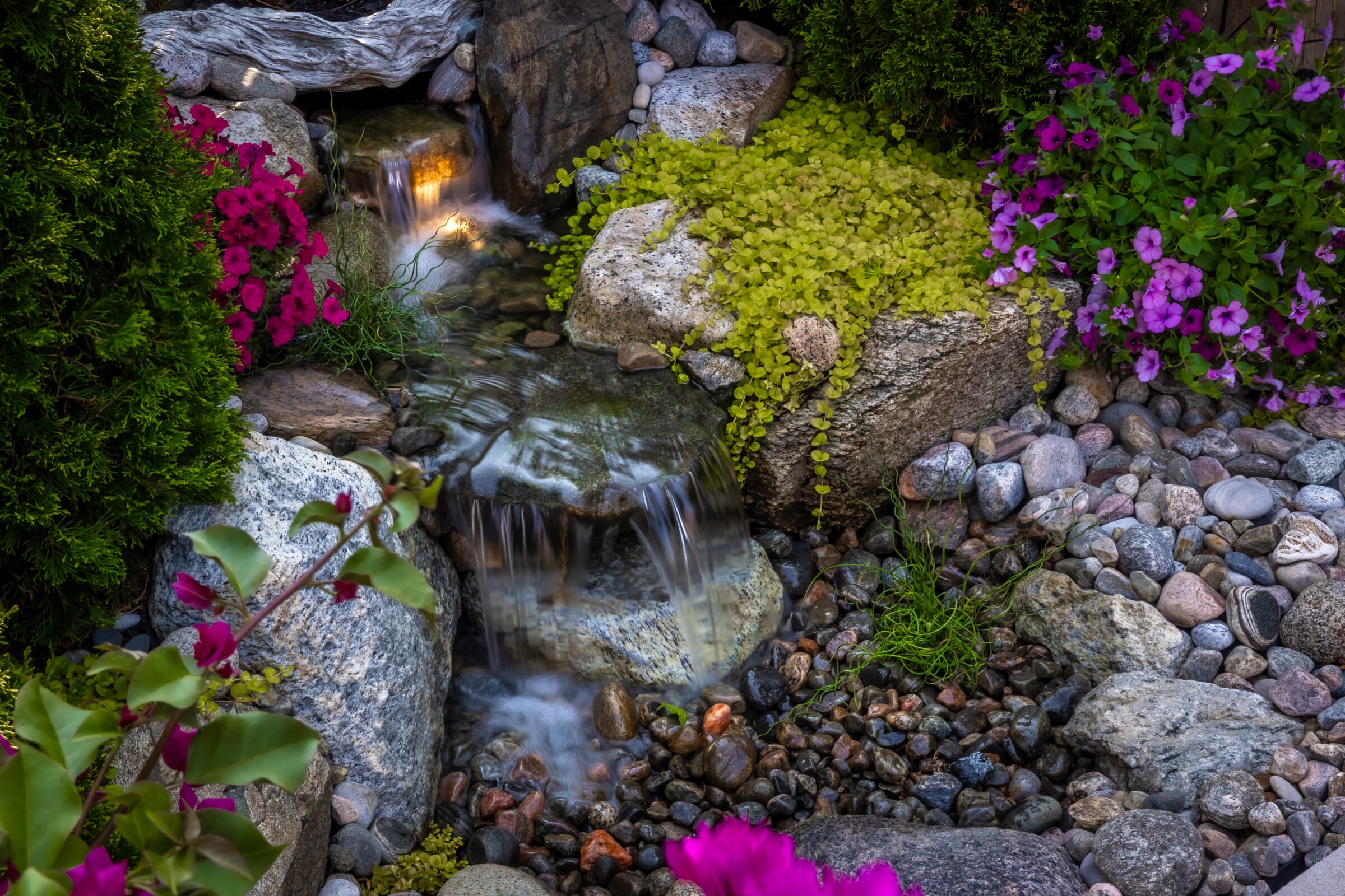 A small garden waterfall flows over rocks, surrounded by vibrant pink and purple flowers, with lush greenery creating a serene atmosphere.