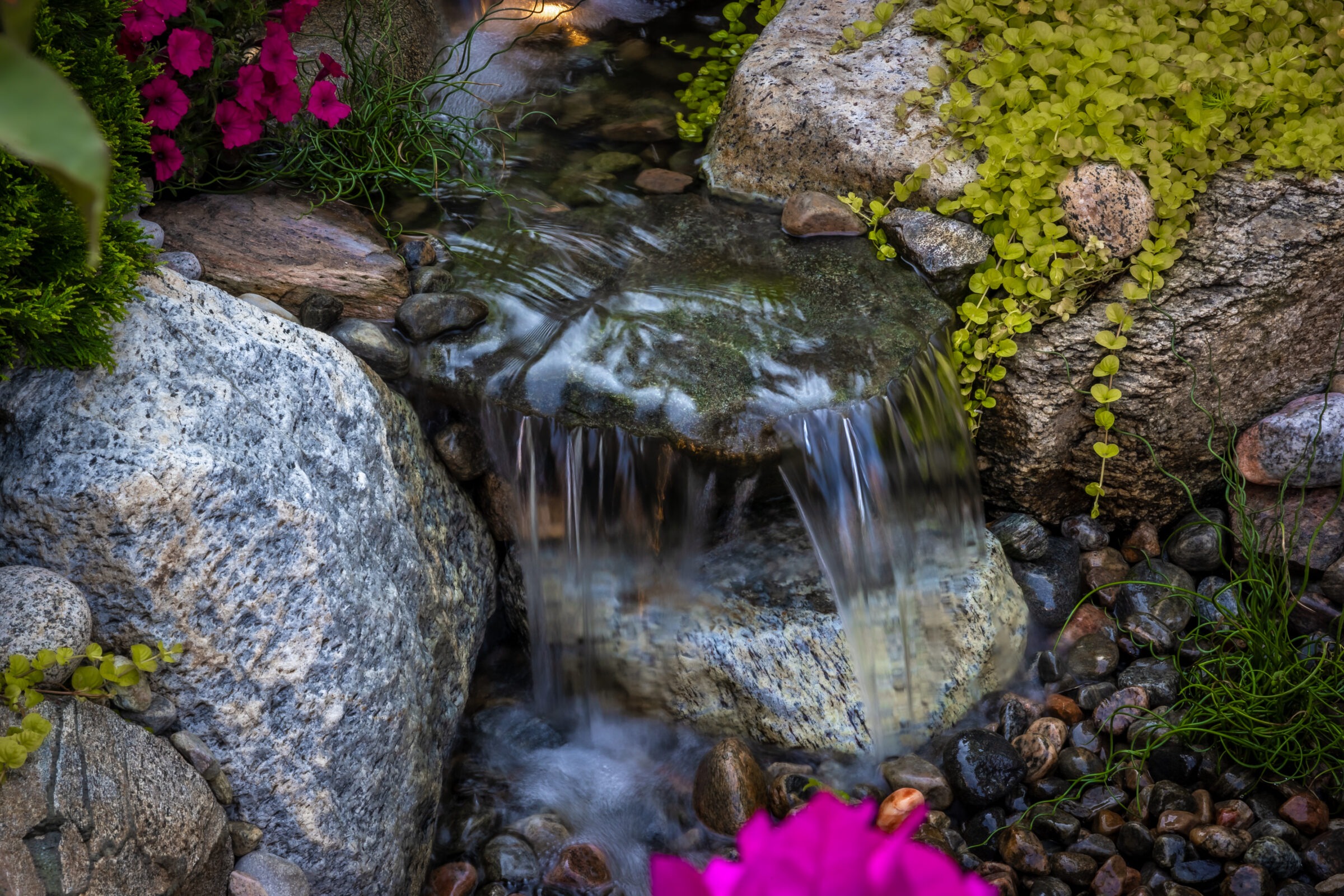 A small, serene waterfall flows over rocks, surrounded by lush greenery and vibrant pink flowers, creating a tranquil garden scene.
