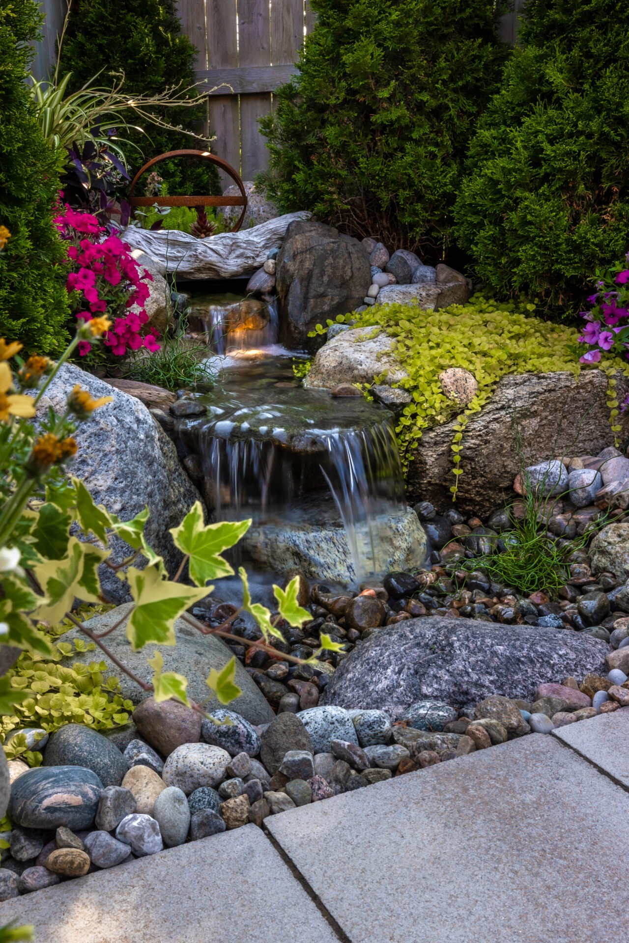 A garden features a small waterfall with stones, lush green plants, and colorful flowers, creating a peaceful, serene atmosphere.
