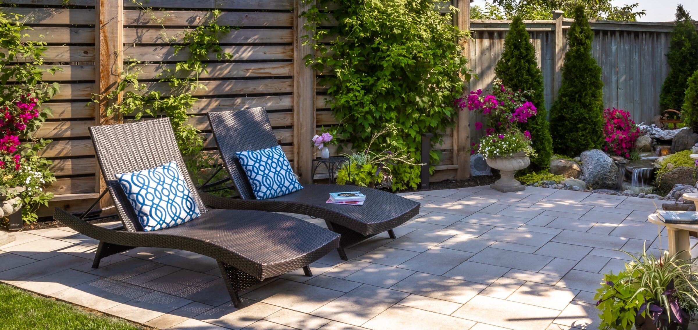 Two wicker lounge chairs with patterned cushions are on a patio surrounded by lush green plants, vibrant flowers, and a small waterfall.