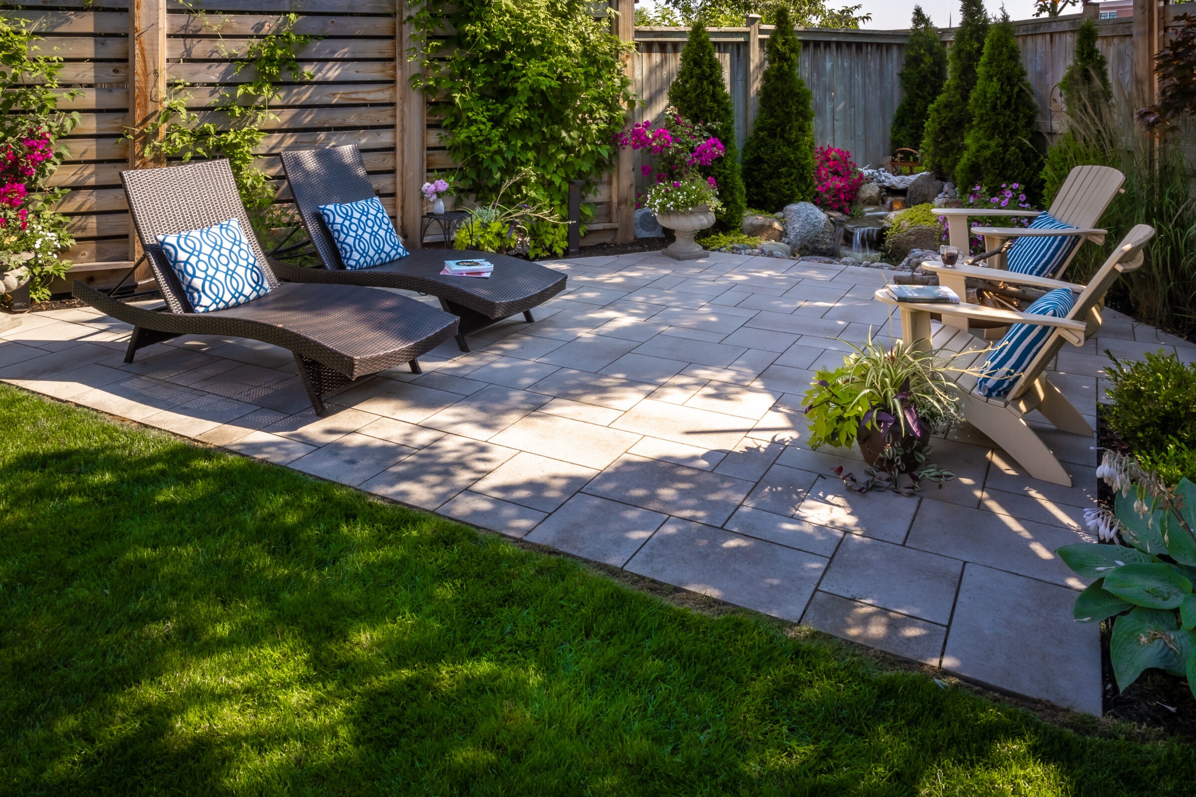 Cozy patio with wicker lounge chairs, wooden Adirondack chairs, vibrant flowers, lush greenery, and a tranquil water feature, on a sunny afternoon.