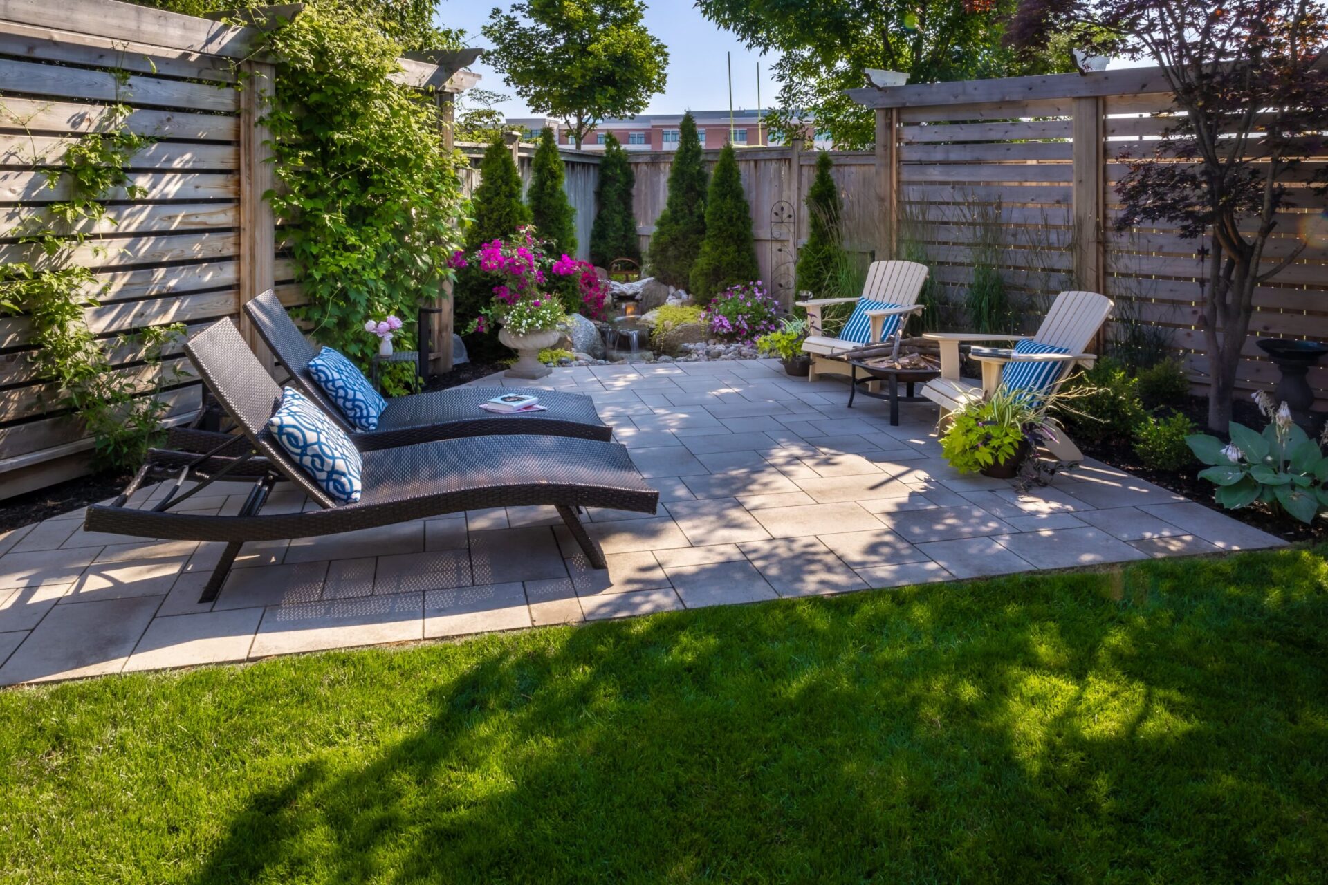 A serene backyard features lounge chairs, vibrant flowers, Adirondack chairs, and a small water feature, all surrounded by wooden fencing and lush greenery.