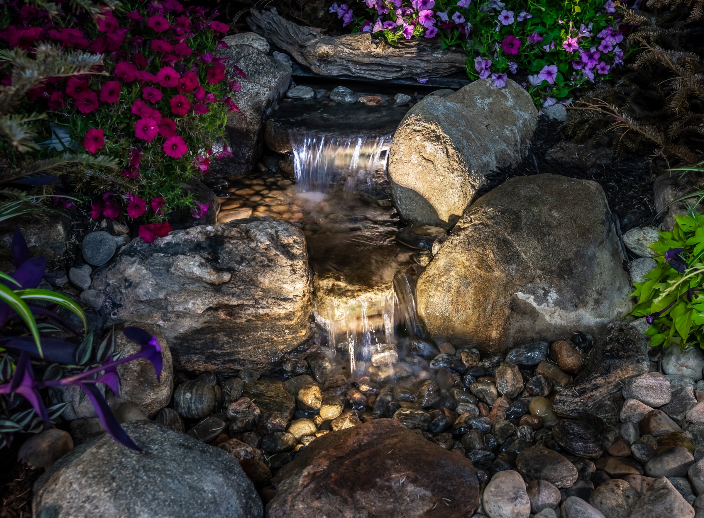 A small waterfall flows over stones, surrounded by vibrant flowers and foliage. Soft lighting highlights the tranquil, natural garden setting.