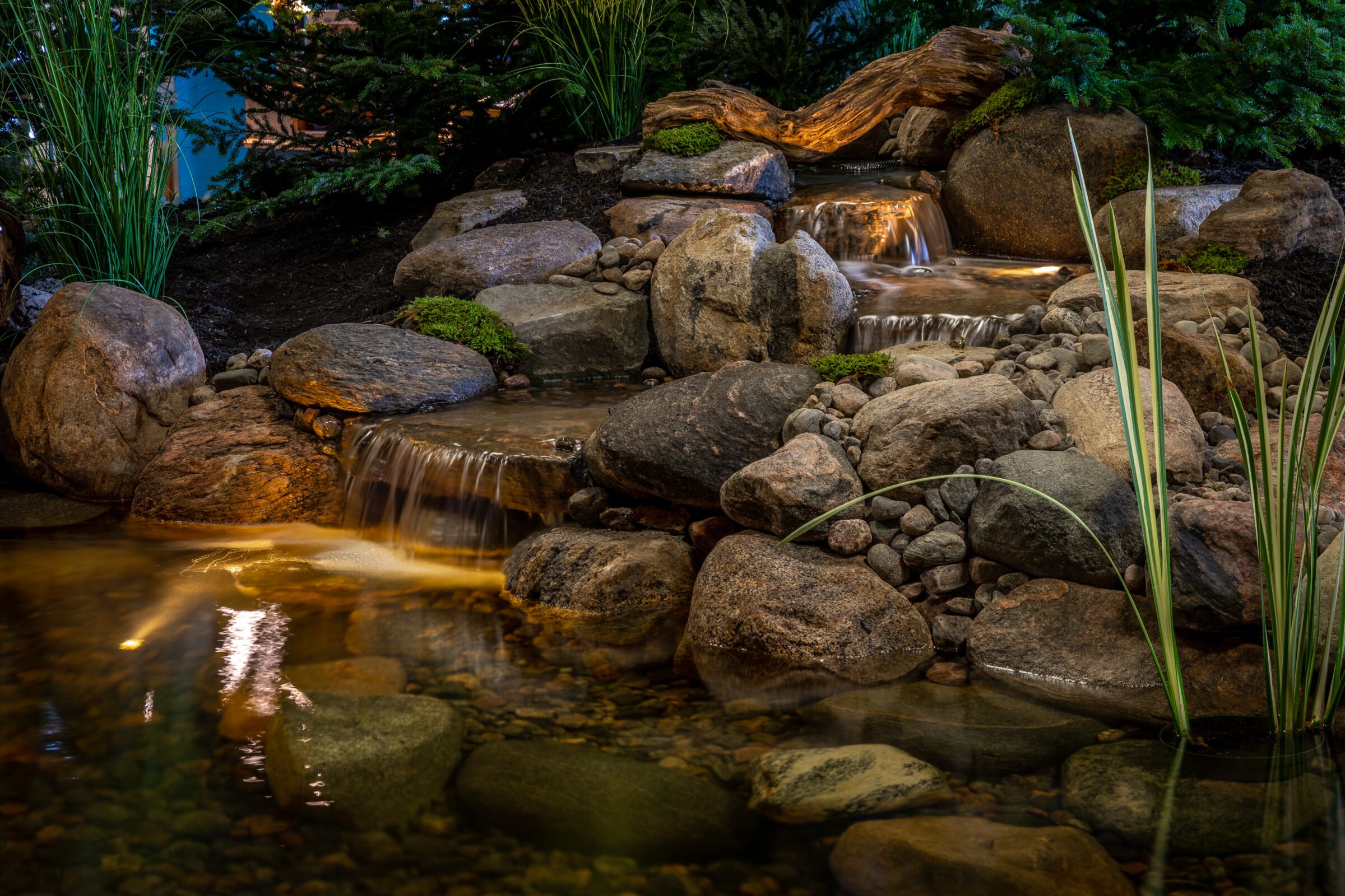 A serene garden scene with a small waterfall cascading over rocks into a pond, surrounded by greenery and illuminated subtly.