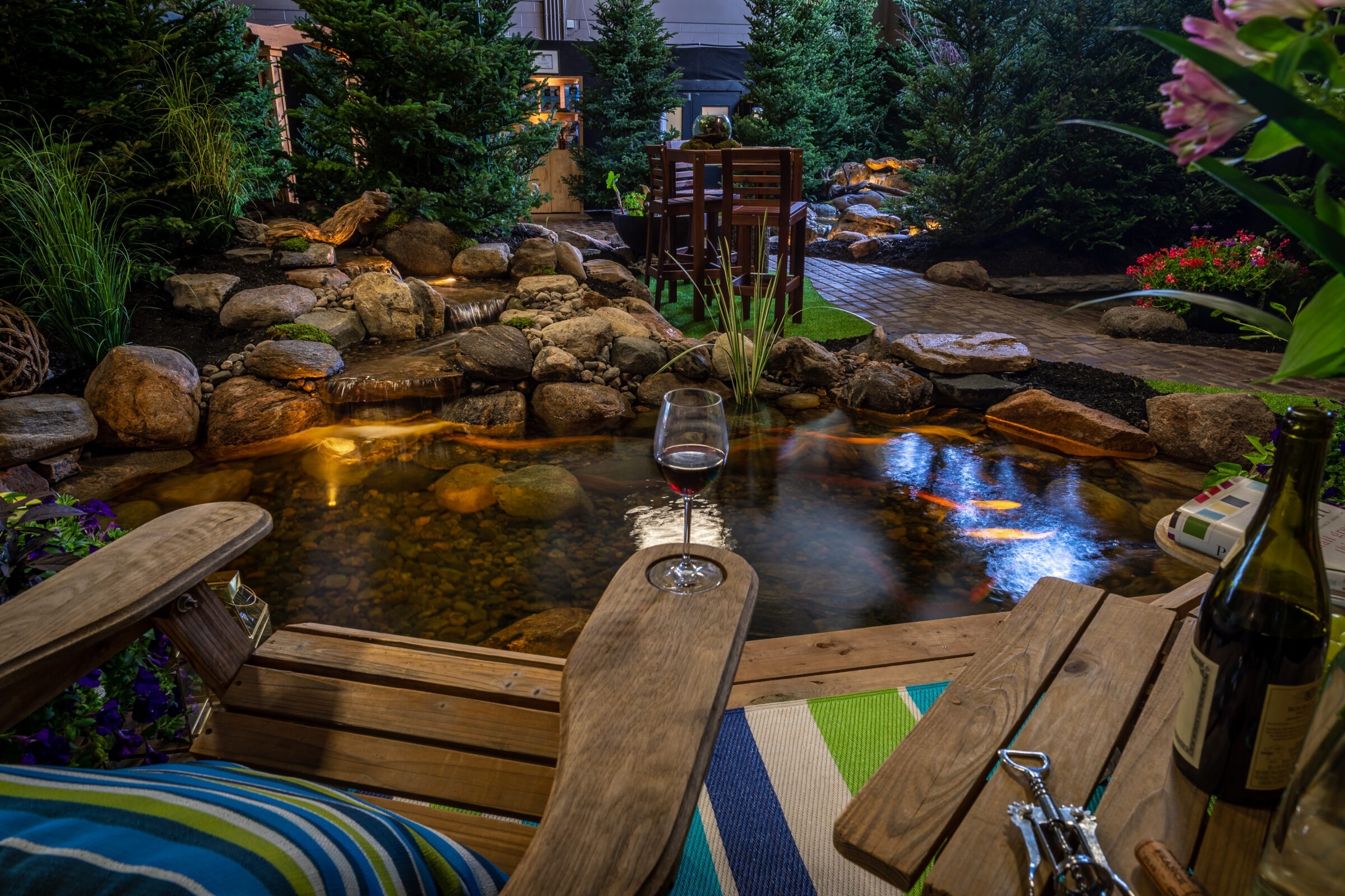 A serene garden setting features a pond, furniture, and wine, surrounded by lush greenery and ambient lighting, creating a peaceful, inviting atmosphere.