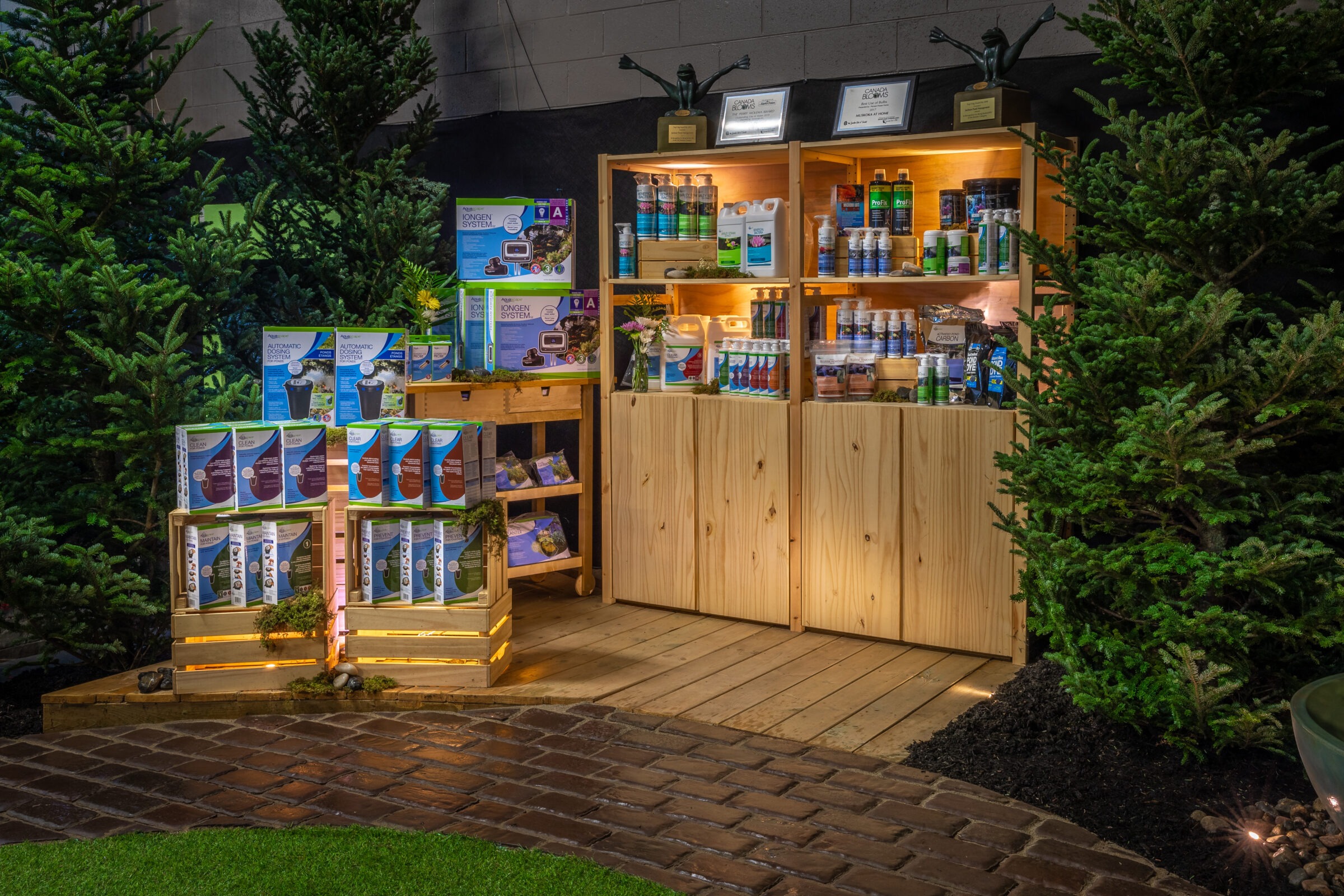 Wooden shelves display garden products, surrounded by pine trees on a brick floor. Lit from above, the setup has a decorative, natural theme.