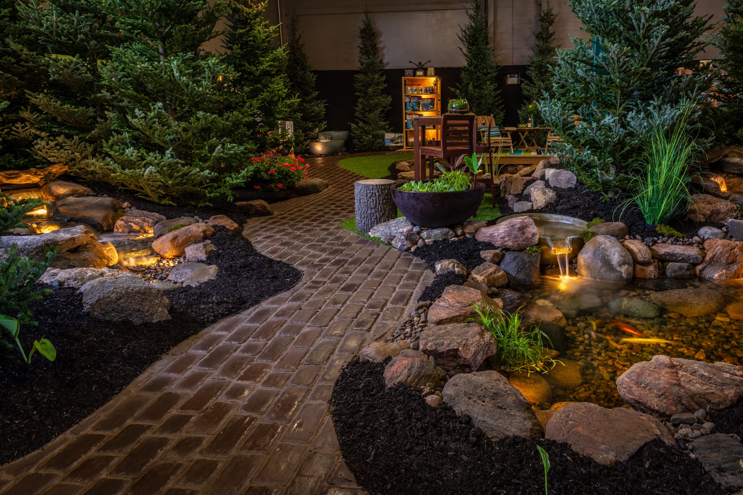 Cozy indoor garden scene with a stone path, illuminated pond, pine trees, wooden furniture, and decorative plants creating a tranquil atmosphere.