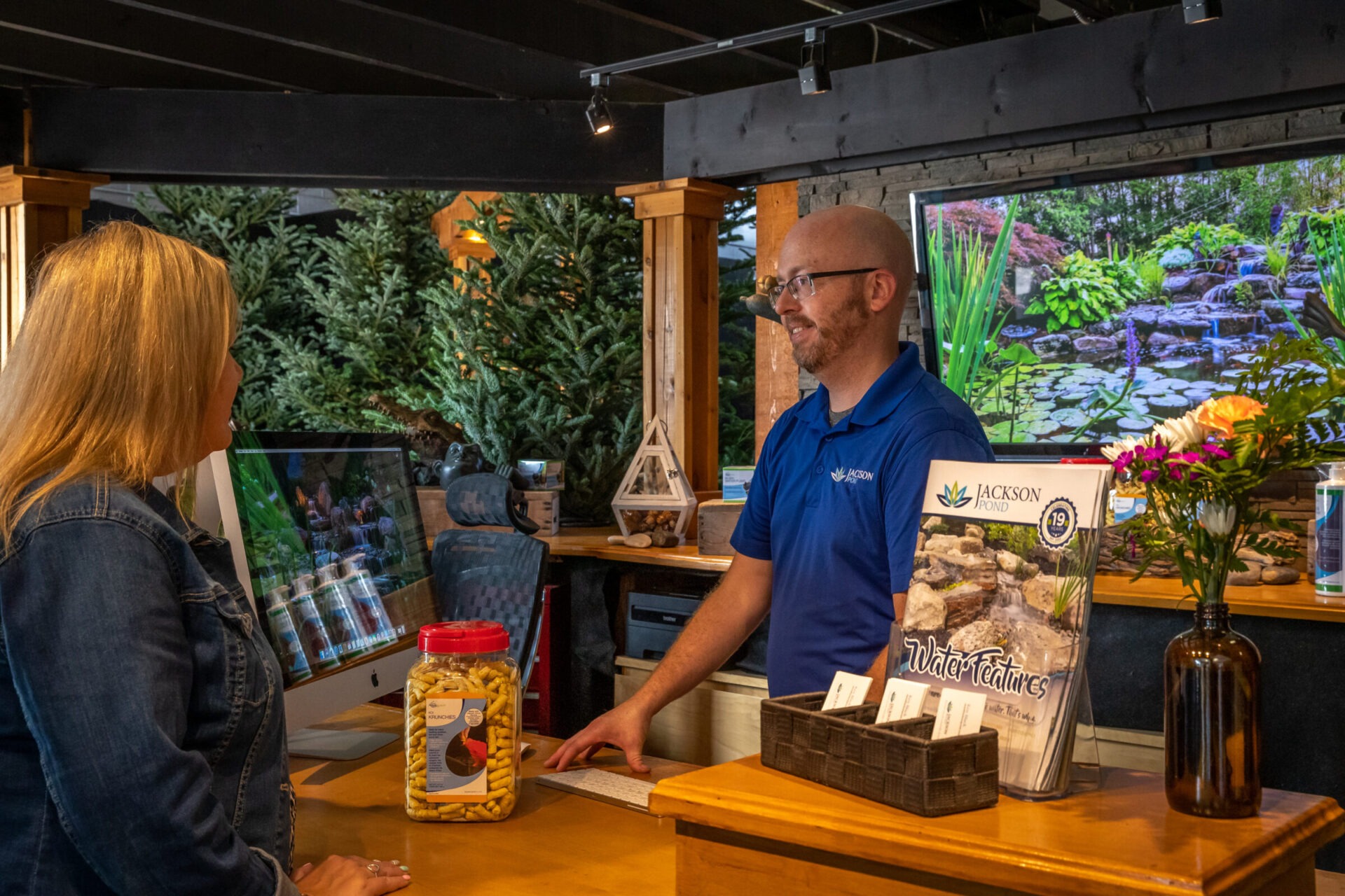 Two people are talking at a reception desk surrounded by greenery, brochures, and a computer displaying a garden scene.