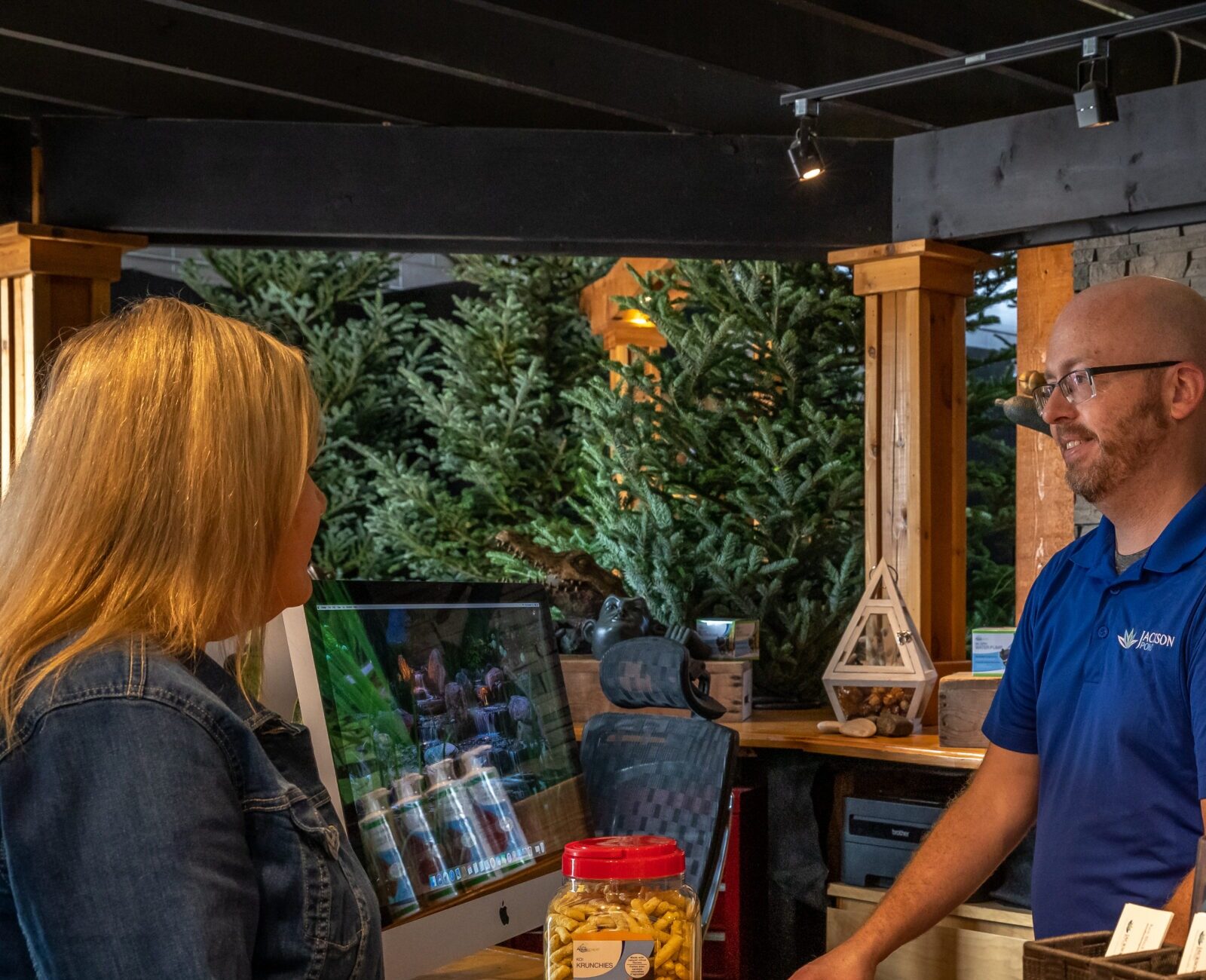 Two people are talking at a reception desk surrounded by greenery, brochures, and a computer displaying a garden scene.