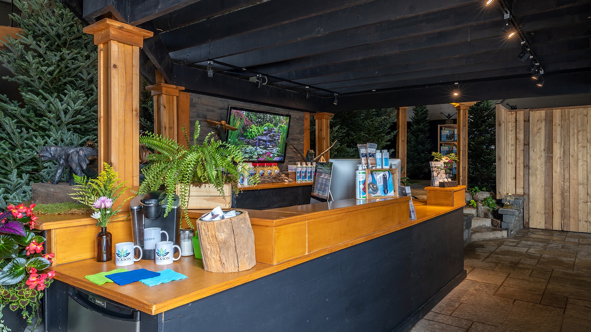 A rustic coffee counter with plants, mugs, and snacks, nestled among evergreens and wood decor, creating a natural, cozy atmosphere.