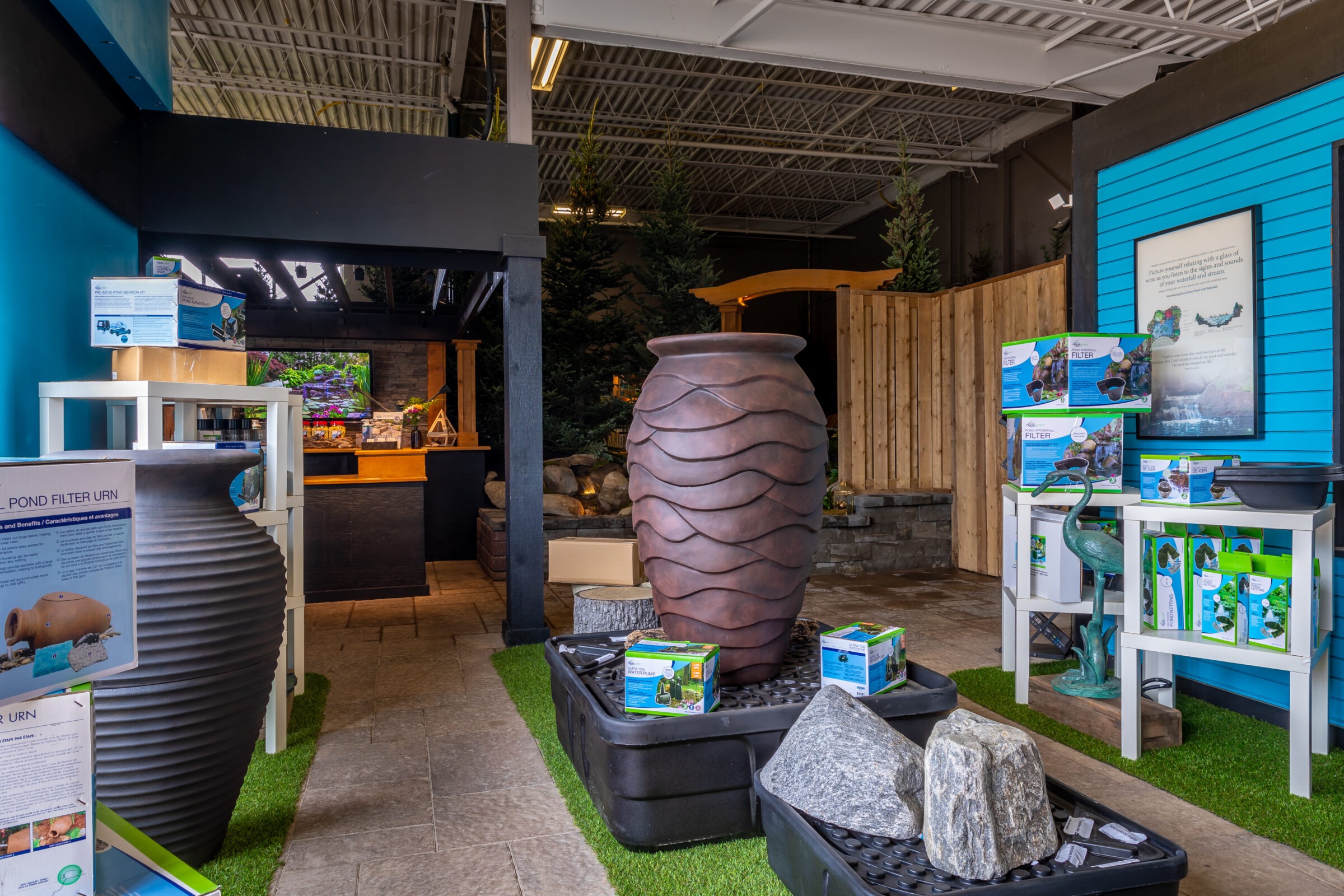 Indoor garden decor shop featuring a large decorative urn, artificial grass, rock displays, and pond filter products, with wooden dividers and vibrant colors.