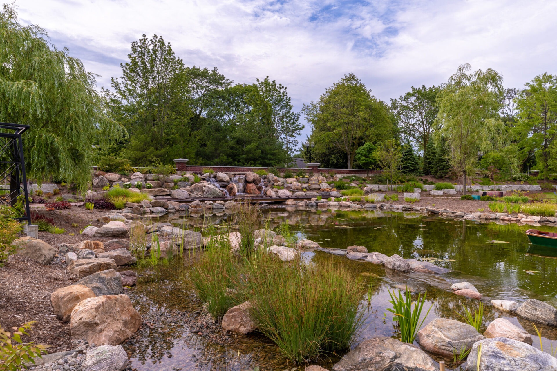 Serene garden scene with a rocky pond, lush greenery, and a small waterfall under a cloudy sky. Peaceful and picturesque setting.