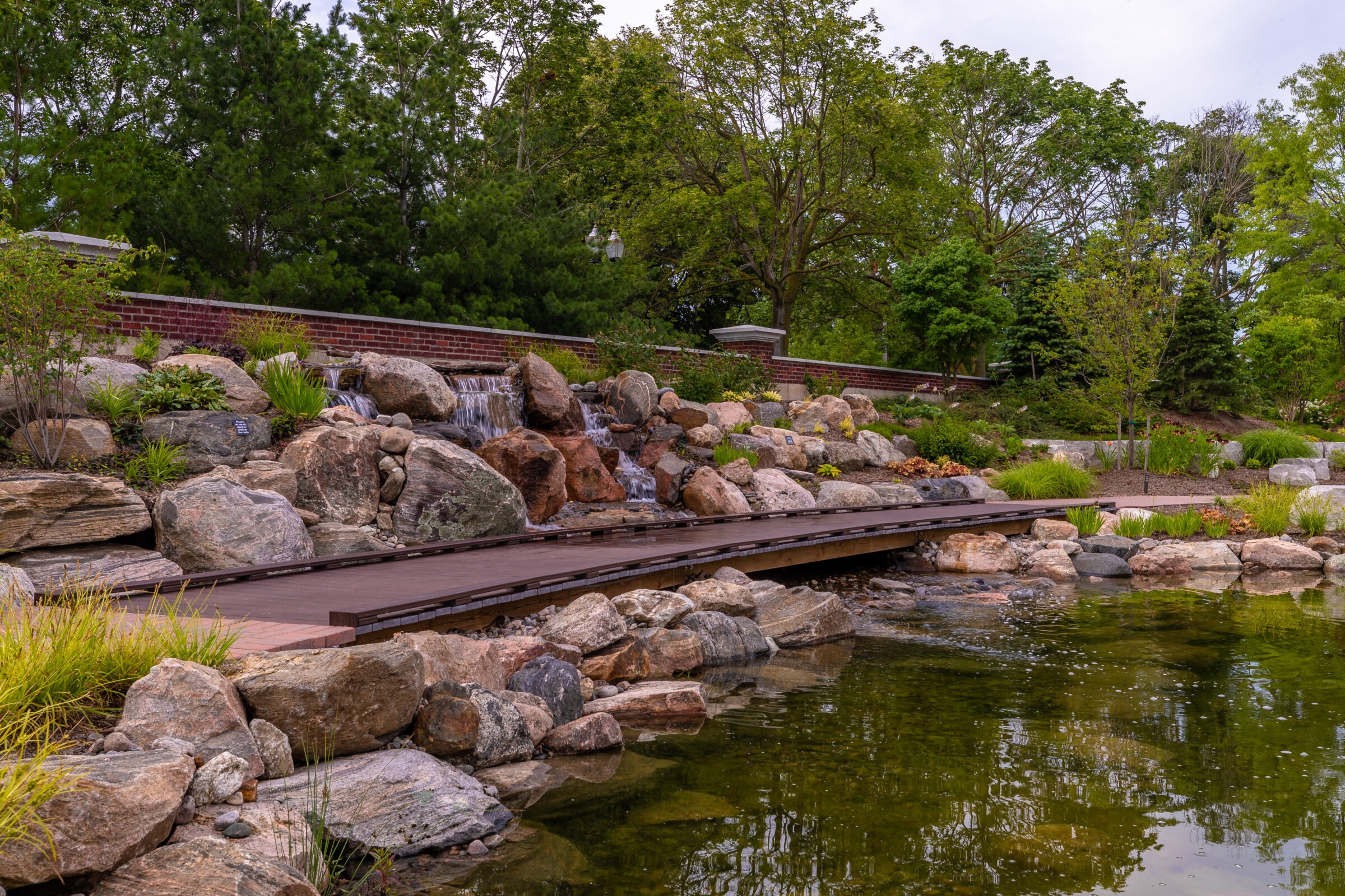A serene garden with a wooden path and cascading waterfall, framed by lush trees and greenery, reflecting in a calm pond.