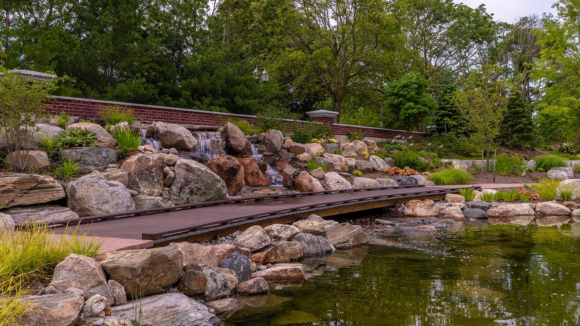 A tranquil garden pond featuring a wooden bridge and waterfall surrounded by rocks and lush greenery on a peaceful day.