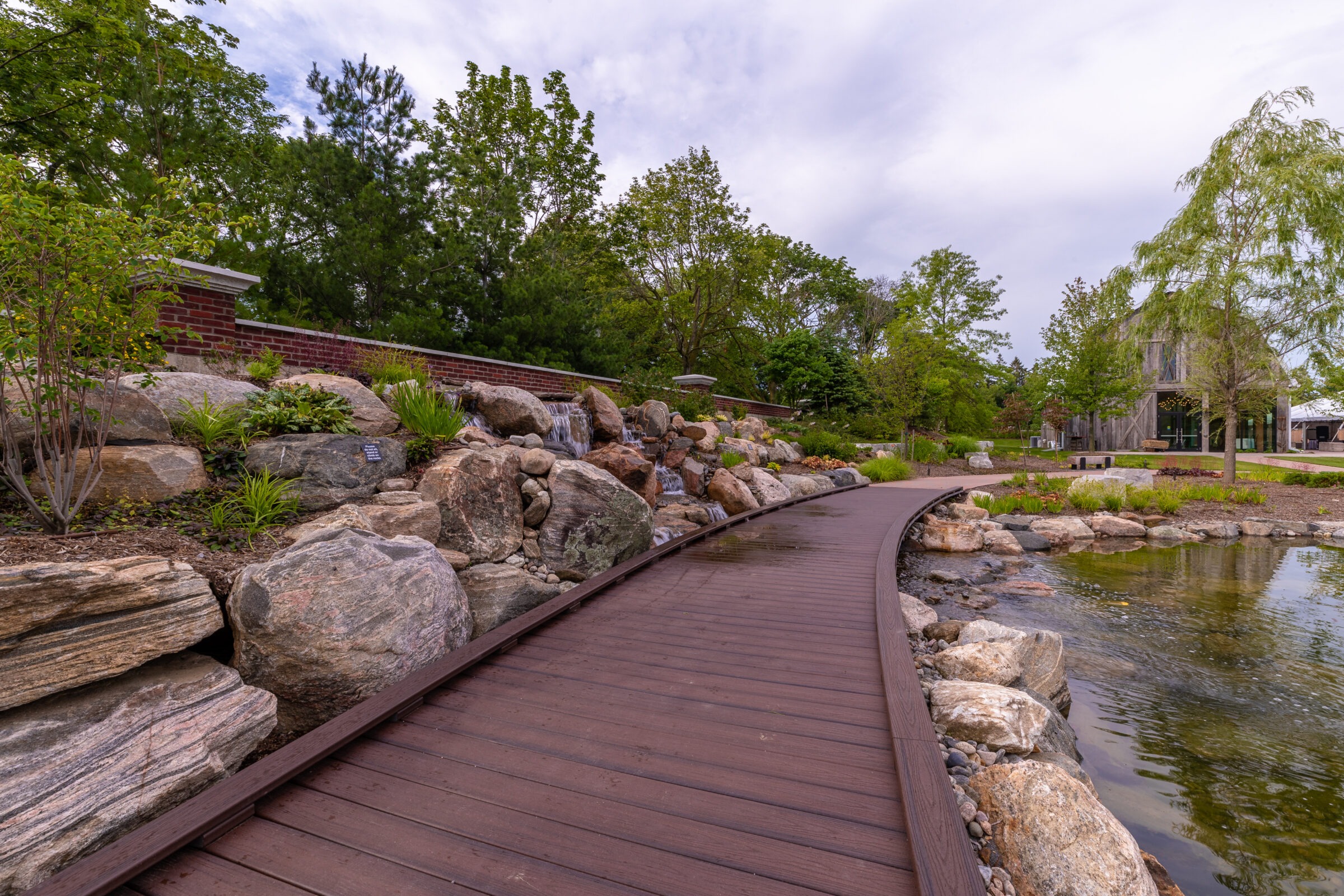 A wooden path beside a pond and rock garden leads to a rustic building, surrounded by lush greenery and a serene atmosphere.