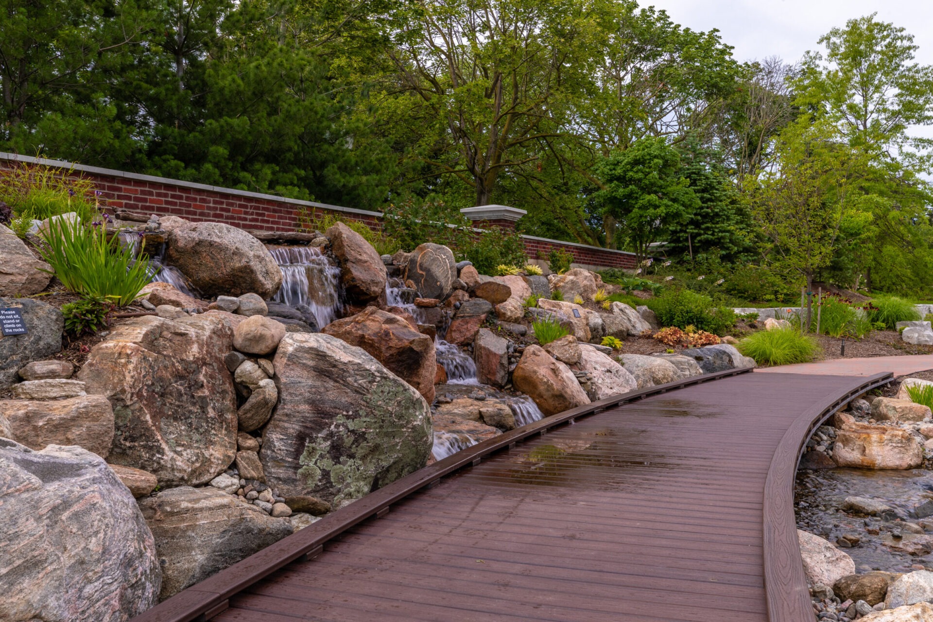 A peaceful garden scene with a wooden walkway, small waterfall over rocks, and lush greenery. A brick wall enhances the serene landscape.