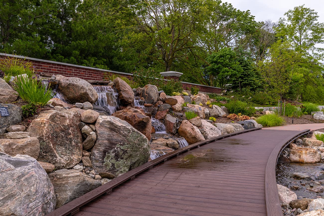 A serene garden path winds alongside a small waterfall over rocky terrain, surrounded by lush greenery and a brick wall background.