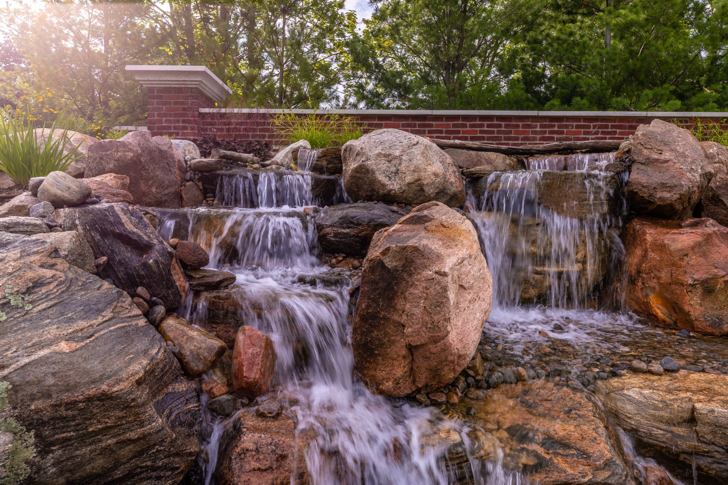 A tranquil scene featuring a small waterfall cascading over rocks, with a brick wall and greenery in the background, under soft sunlight.