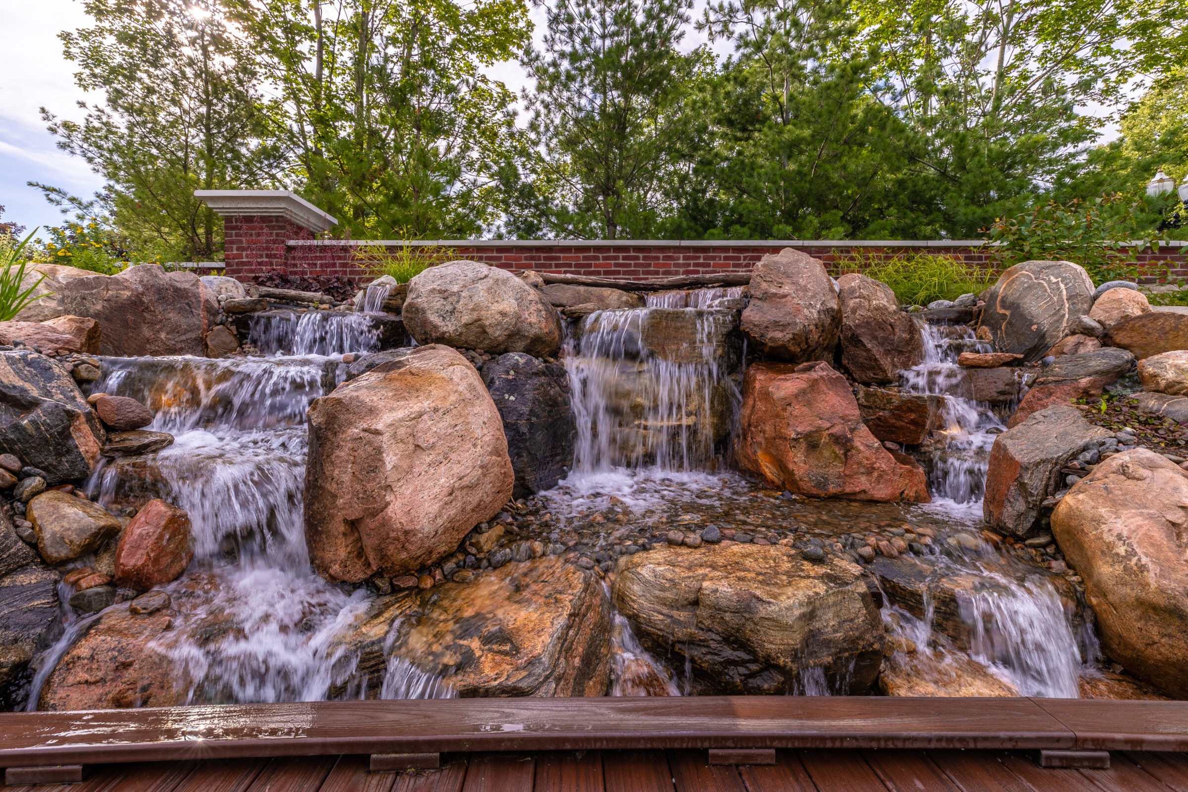 A small waterfall flows over large rocks, surrounded by greenery, a brick wall, and wooden walkway, creating a serene natural setting.