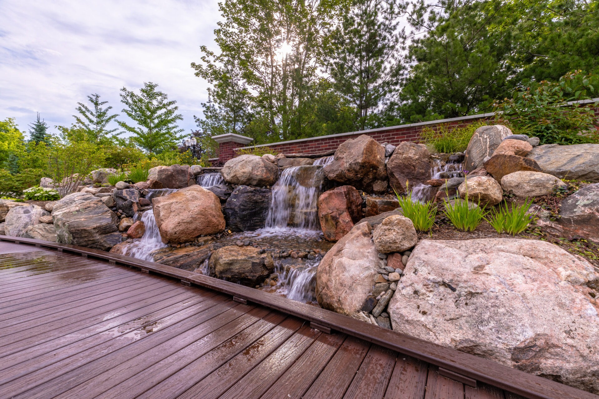 A serene waterfall cascades over rocks in a garden setting, surrounded by trees and a wooden walkway under a partly cloudy sky.