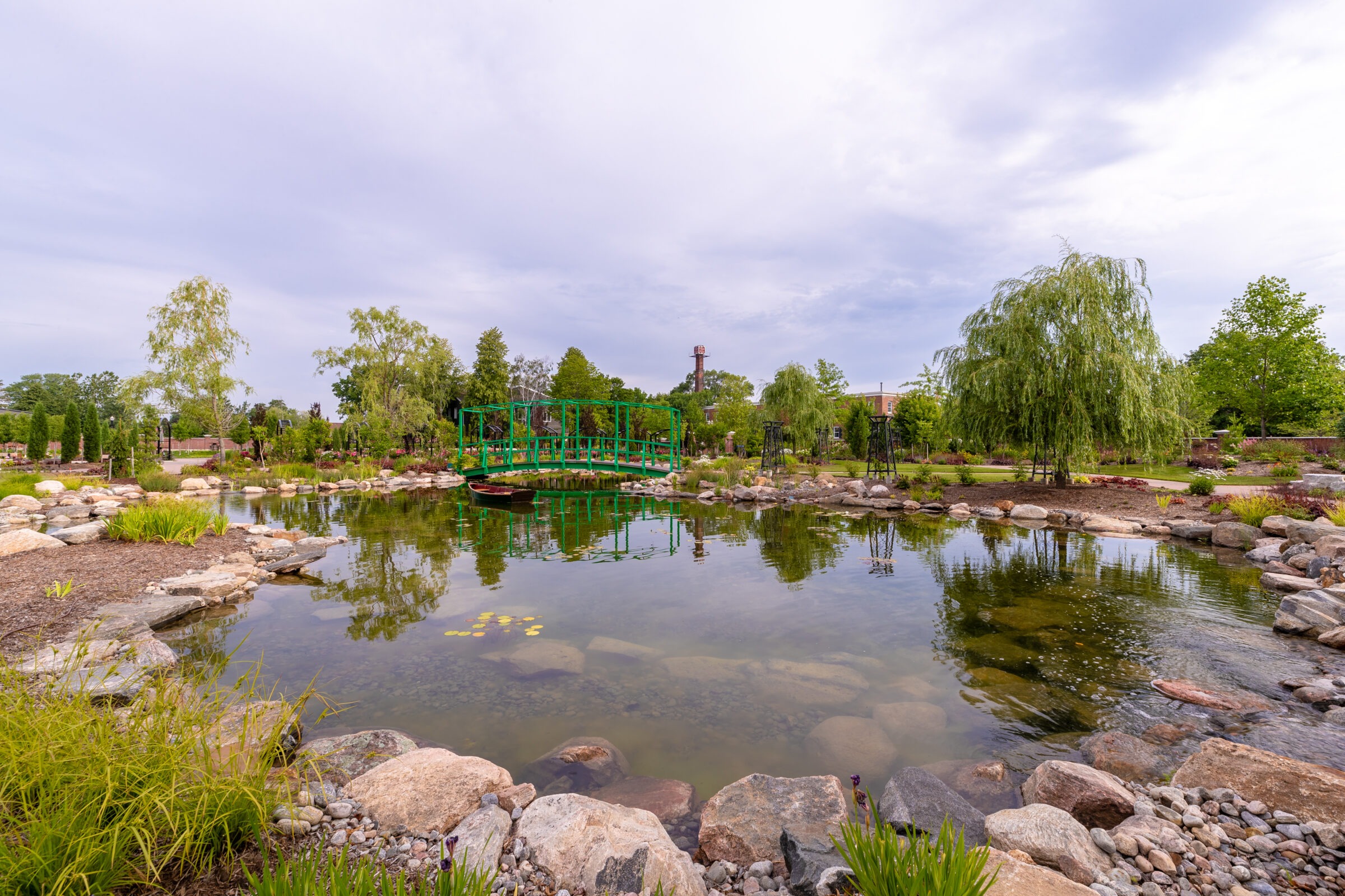 A serene garden scene with a small pond, green bridge, and lush trees under a cloudy sky. Peaceful, natural landscape setting.