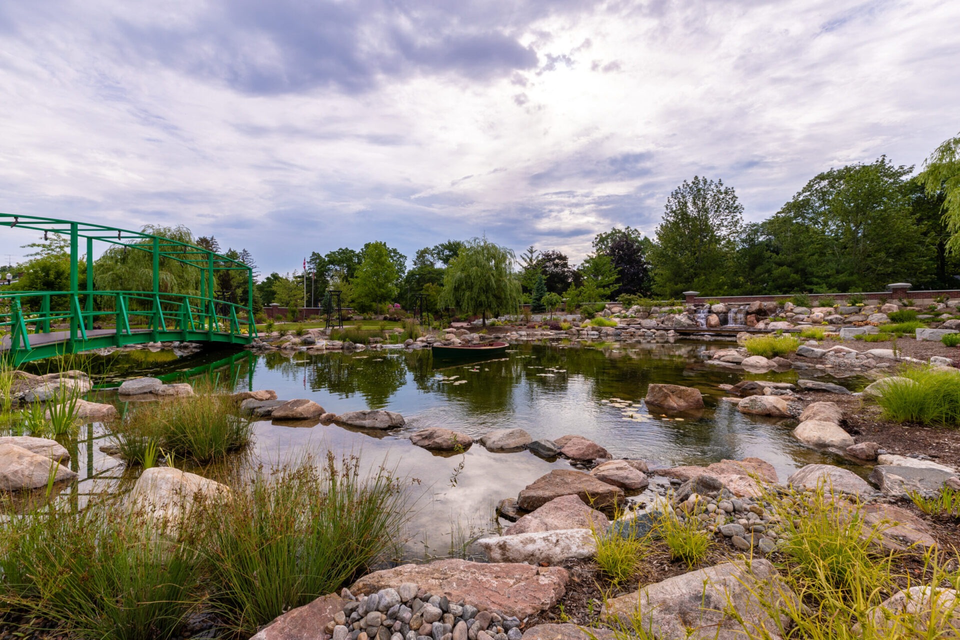 A serene landscape featuring a green bridge, tranquil pond, rocks, lush trees, and vegetation under a cloudy sky. No people visible.