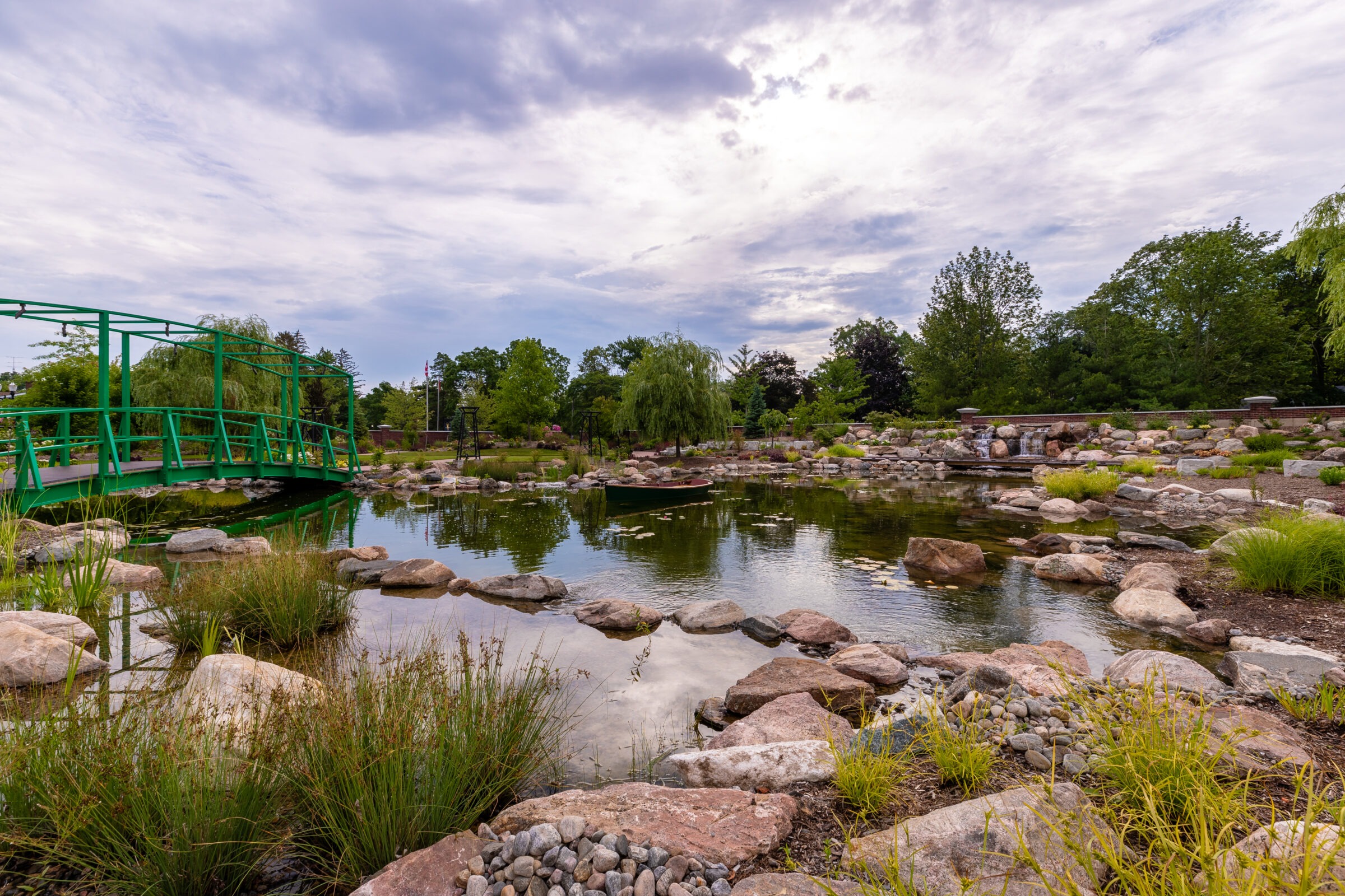 A serene landscape featuring a green bridge, tranquil pond, rocks, lush trees, and vegetation under a cloudy sky. No people visible.