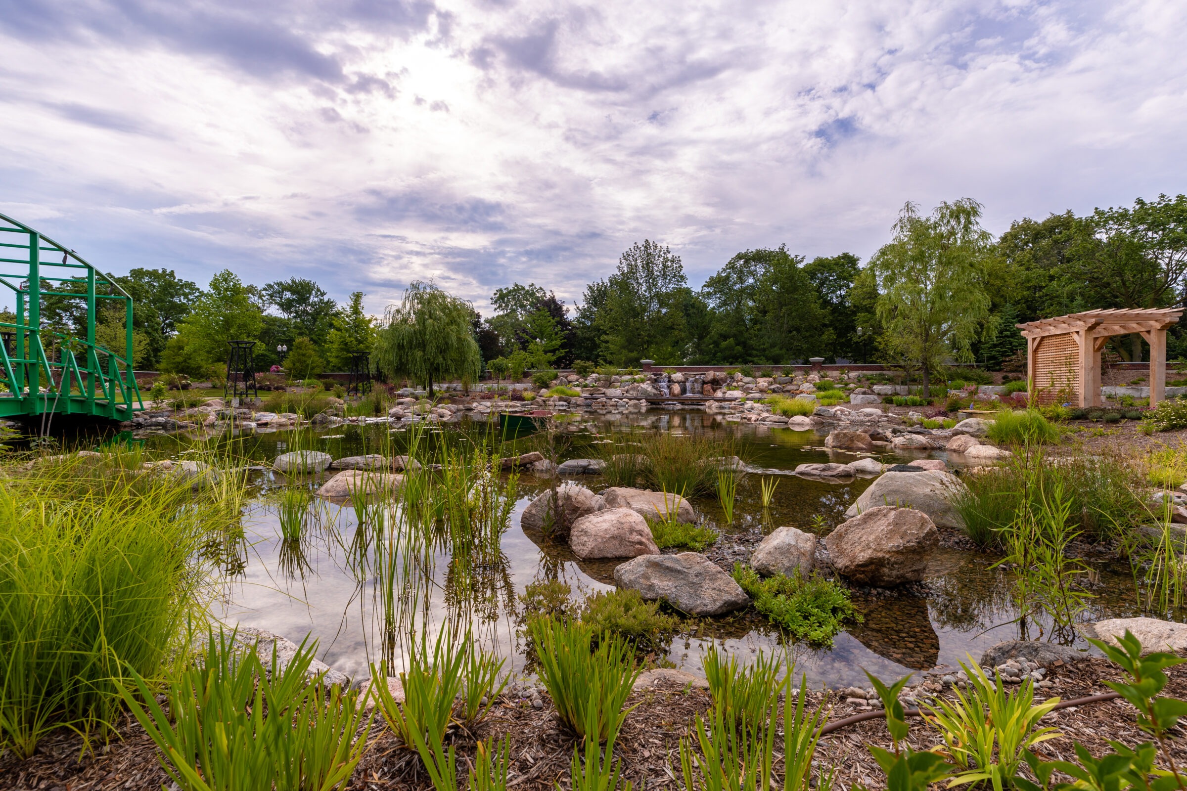 Tranquil garden scene with a green bridge, rocks, and lush vegetation under a cloudy sky. Wooden pergola adds to the serene atmosphere.