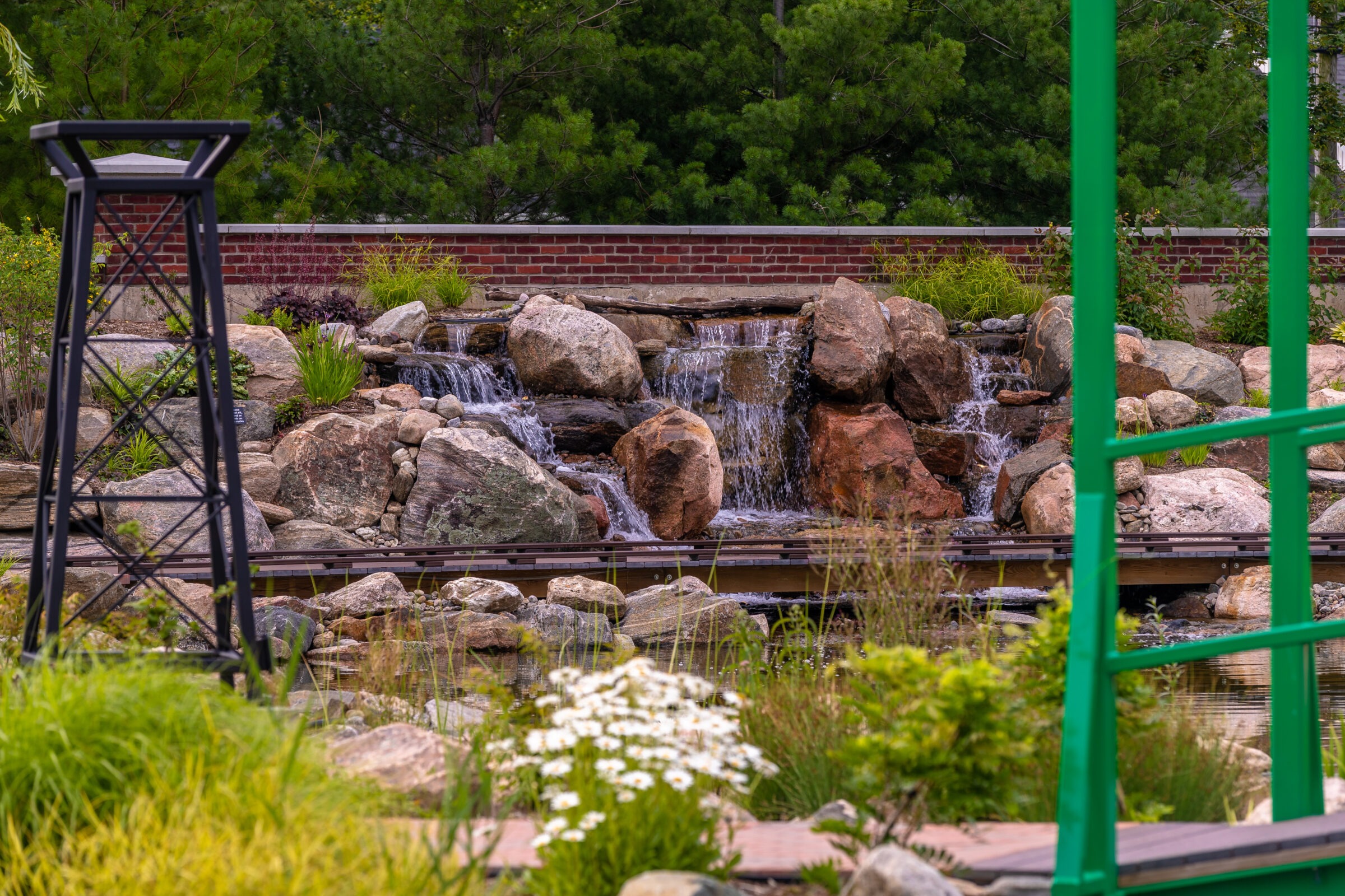 A tranquil garden scene features a small waterfall over rocks, surrounded by lush greenery, flowers, and metal structures, with a brick wall backdrop.