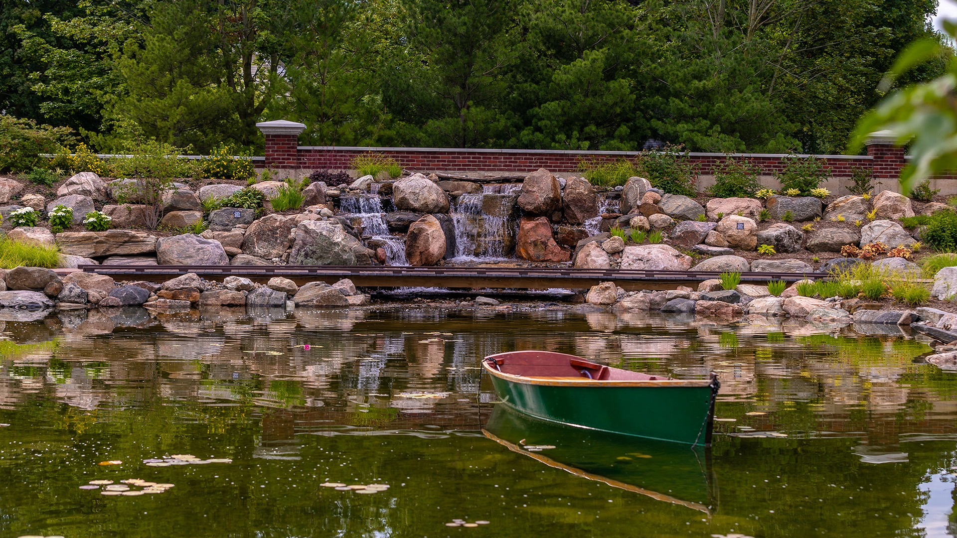 A serene pond features a small waterfall, surrounded by rocks and greenery, with a green boat floating in the foreground.