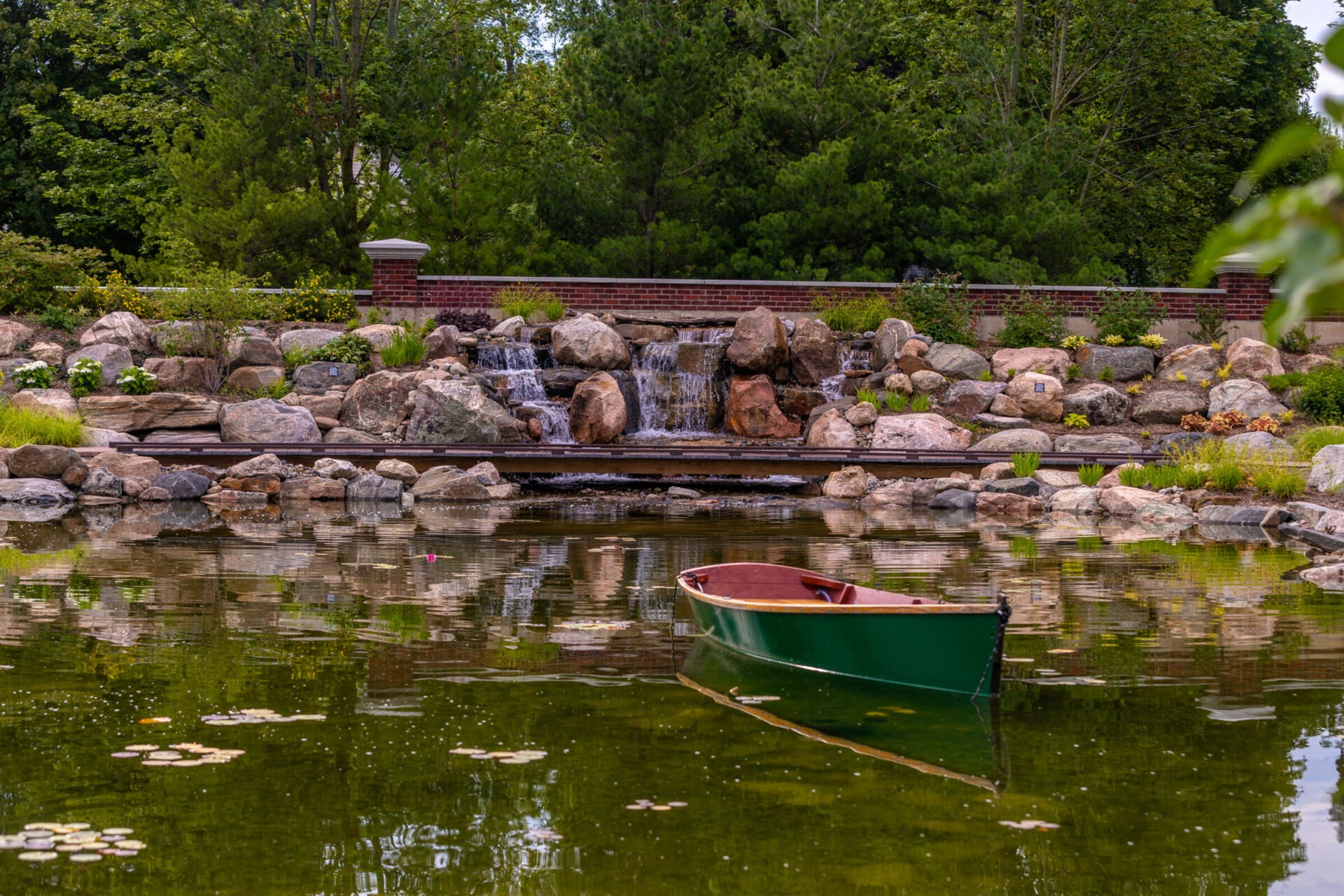 A serene pond scene with a small waterfall, surrounded by rocks and greenery, features an empty green boat floating on the water.