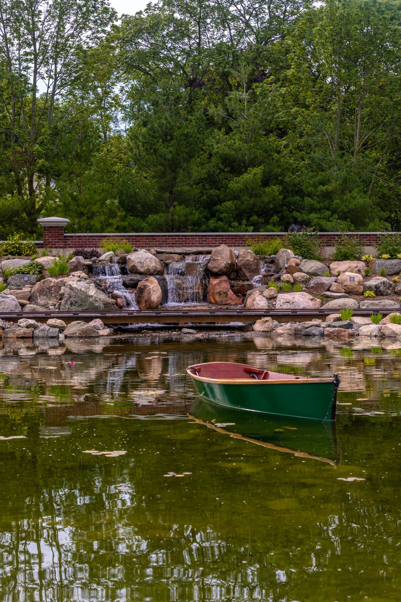 A serene pond with a solitary green boat, surrounded by trees and rocks, featuring a small waterfall and brick accents.