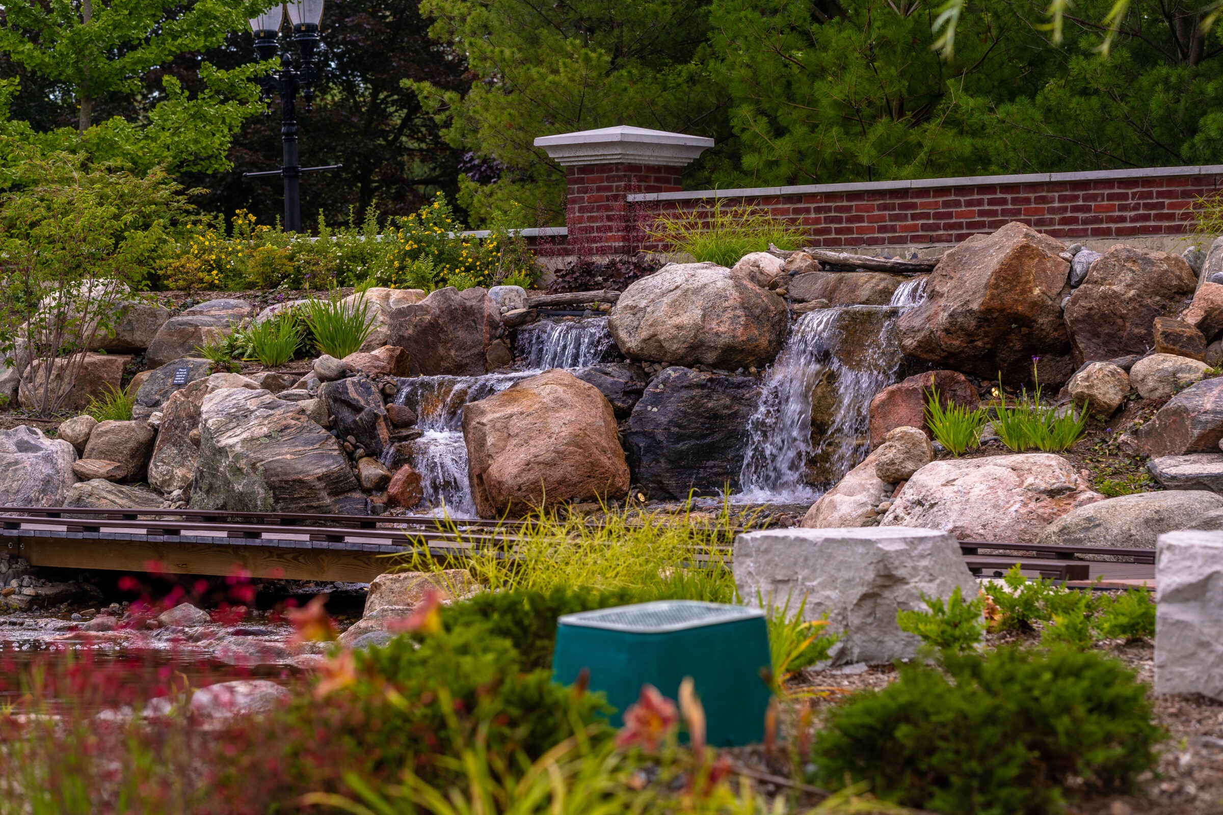 A serene garden scene featuring a small waterfall over rocks, surrounded by greenery and flowers, with a brick wall and lamppost behind.