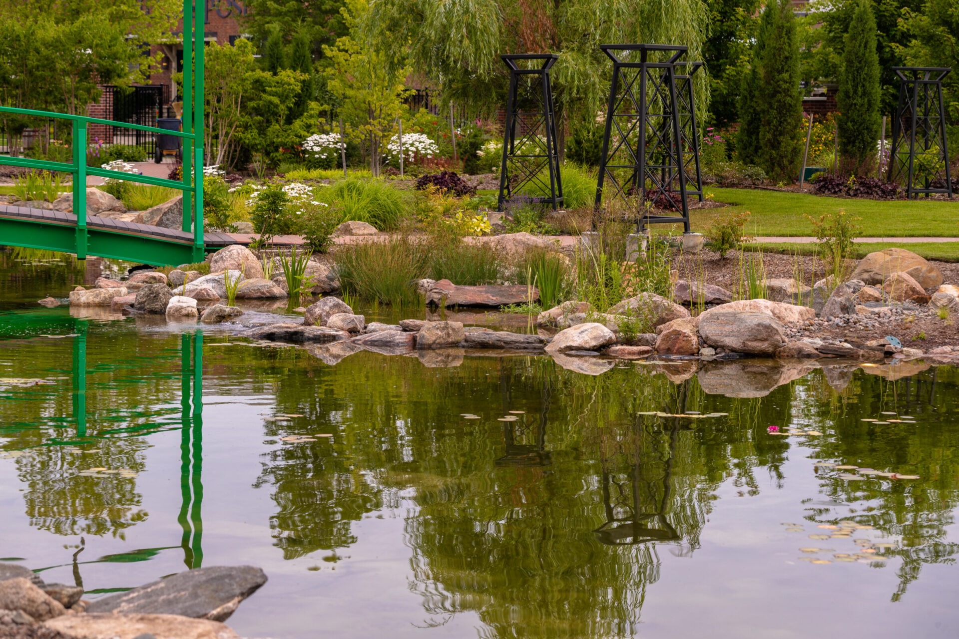 A serene garden features a green bridge over a tranquil pond, surrounded by rocks, plants, and metal structures amidst lush greenery.