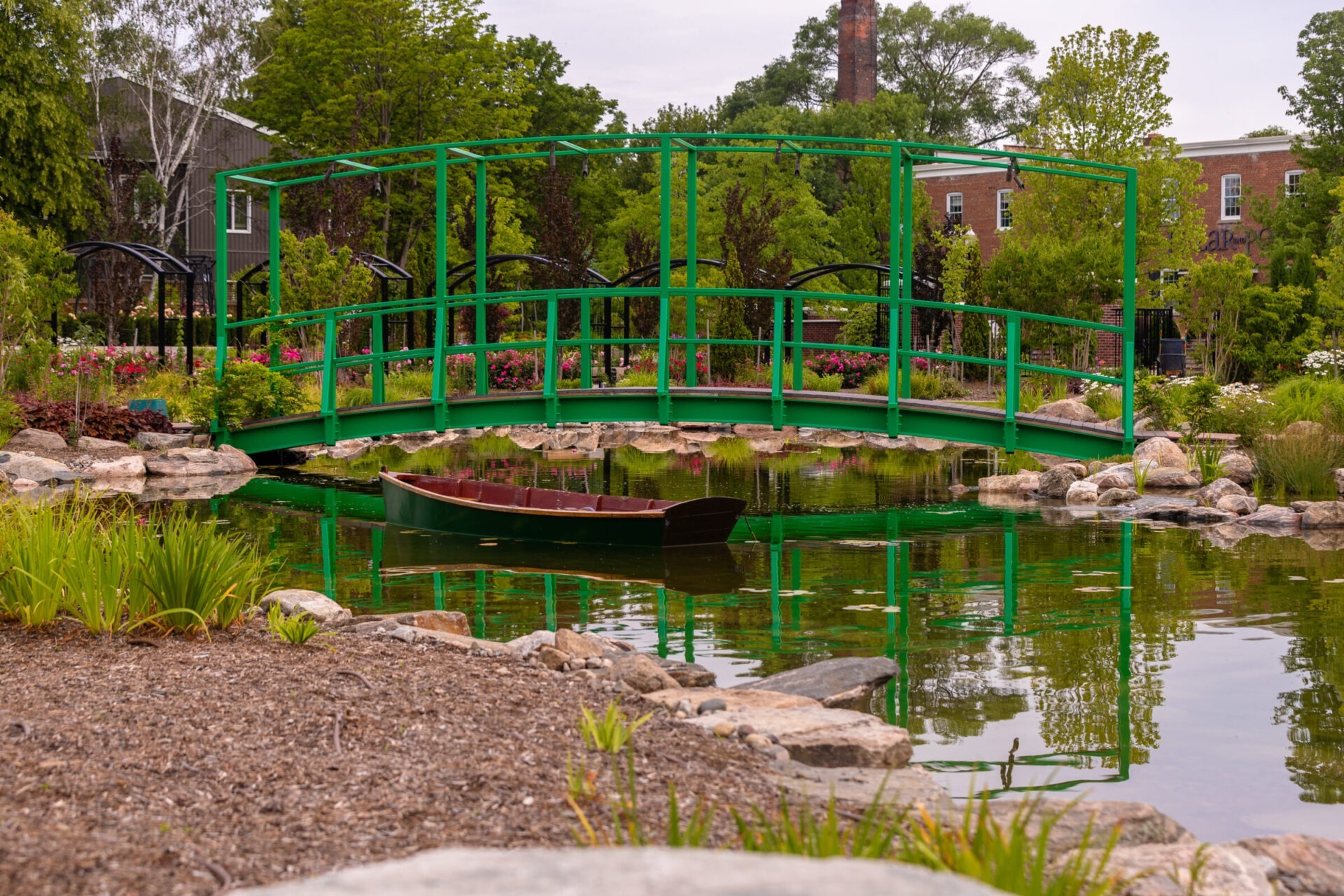 A vibrant green bridge spans a serene pond with a boat, surrounded by lush gardens and a brick building in the background.