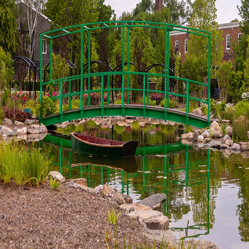 A green bridge spans a tranquil pond with a small boat, surrounded by lush vegetation and a nearby brick building.