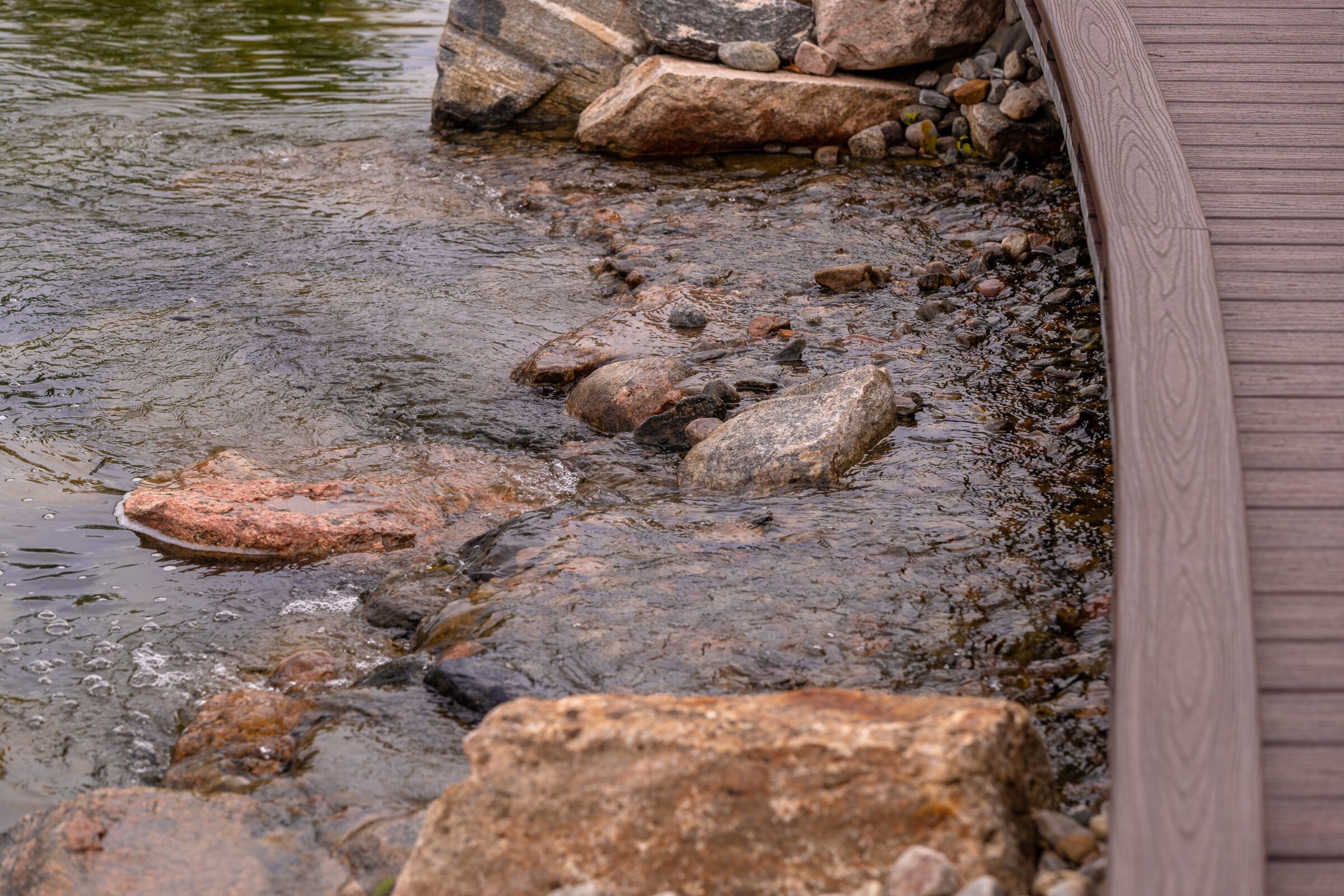Curved wooden deck beside a small stream with smooth rocks, gently flowing water, and lush greenery in the background. Peaceful natural setting.